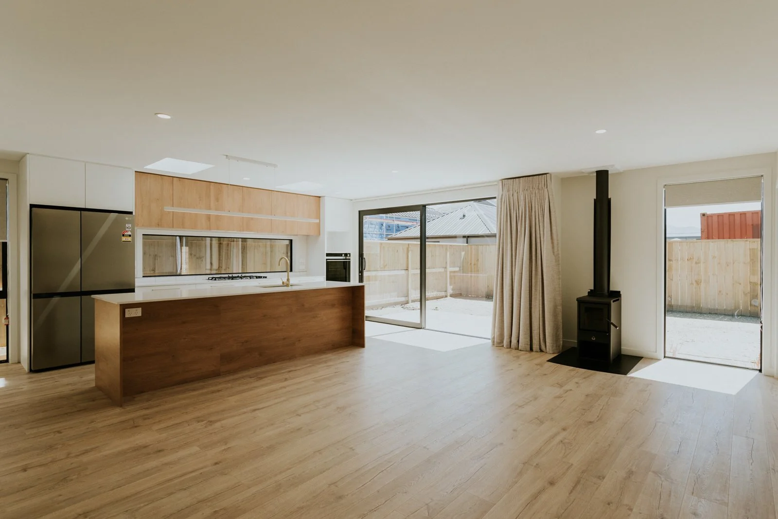 Empty living room with wooden flooring, kitchen area, sliding glass doors leading to backyard, and a wood stove in the corner.