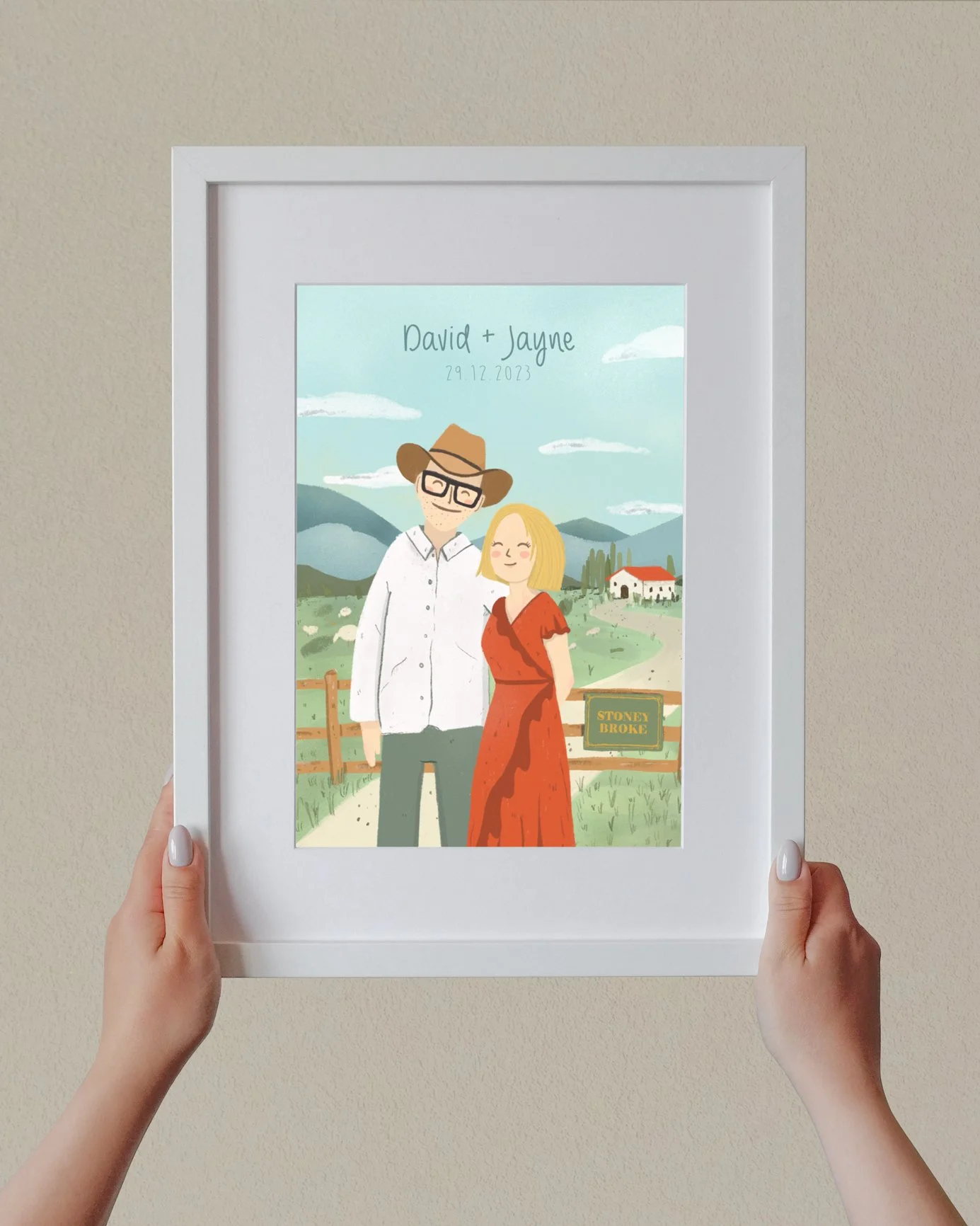 Framed digital illustration of a couple standing outdoors, with the names 'David + Jayne' and the date '29.12.2023' at the top. The background features mountains, cloudy sky, and a house with a road and fencing. A sign reads 'Stoney Broke'.