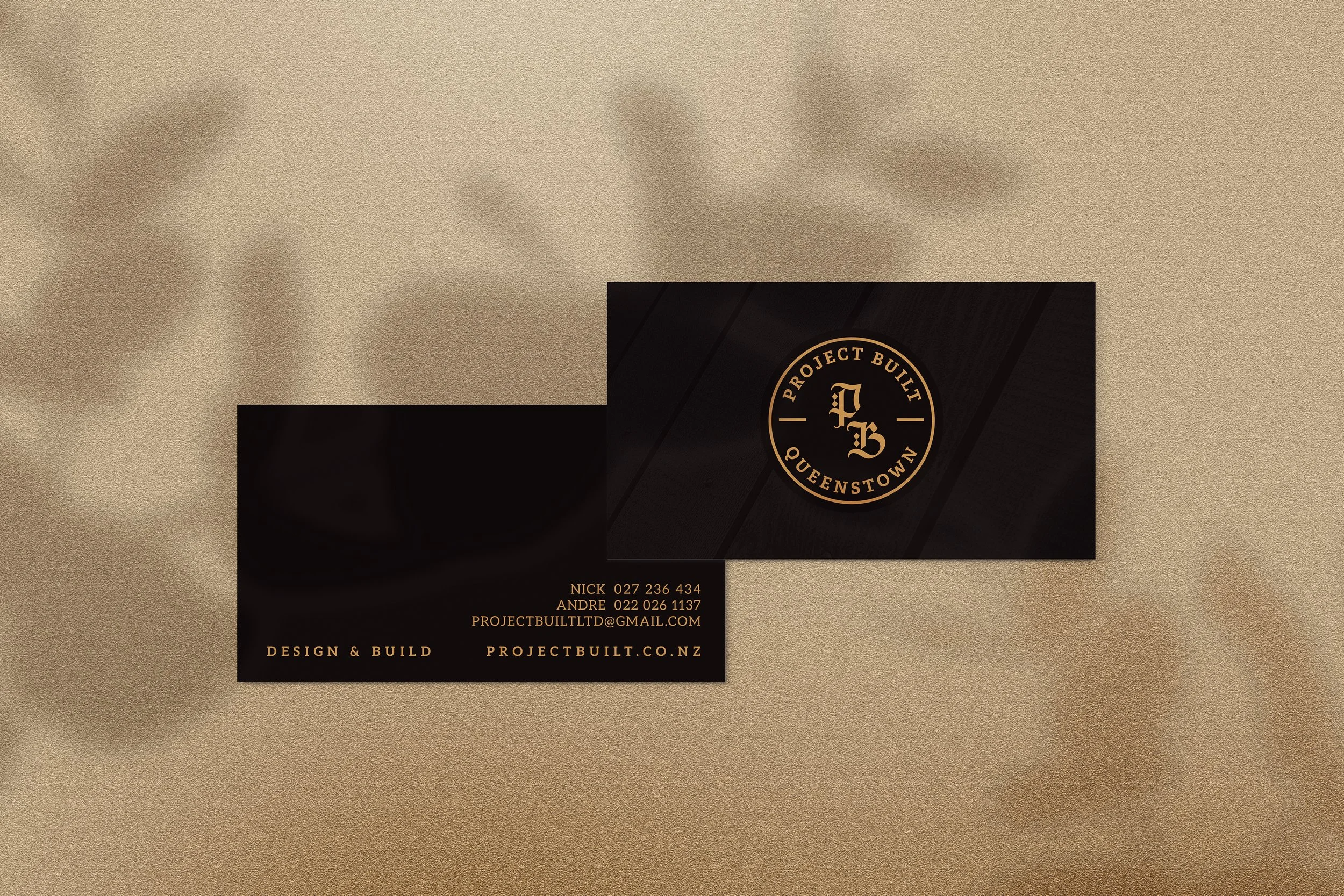 Business cards for Project Built, a construction company based in Queenstown. The cards are black with gold text and logo, placed on a beige textured surface with blurred shadows.