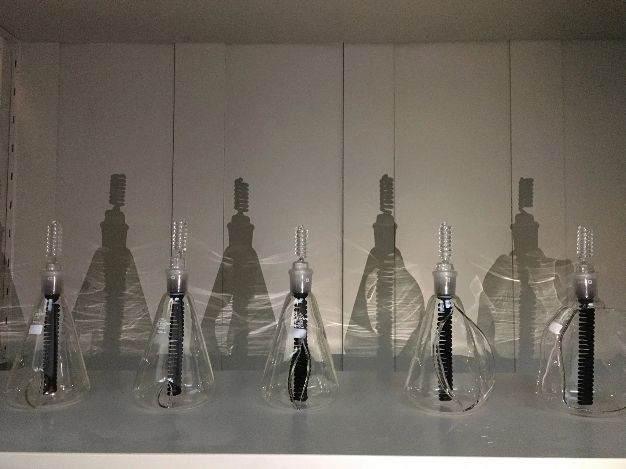 a row of glass flasks with dark glass coils inside and a light casting shadows