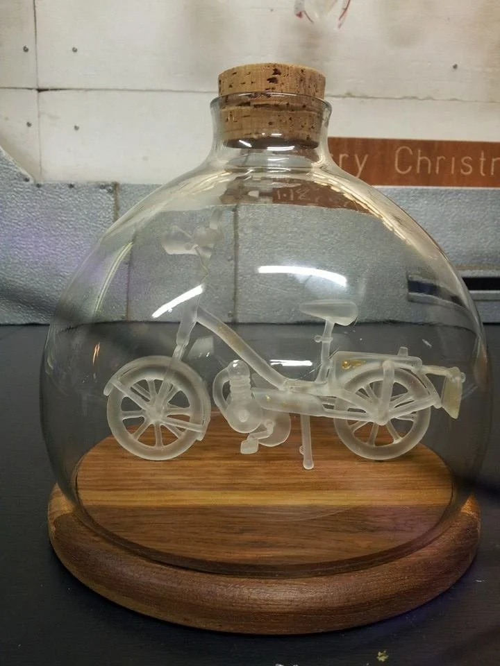 A frosted glass moped inside a wide glass flask with a cork stopper on a wooden base