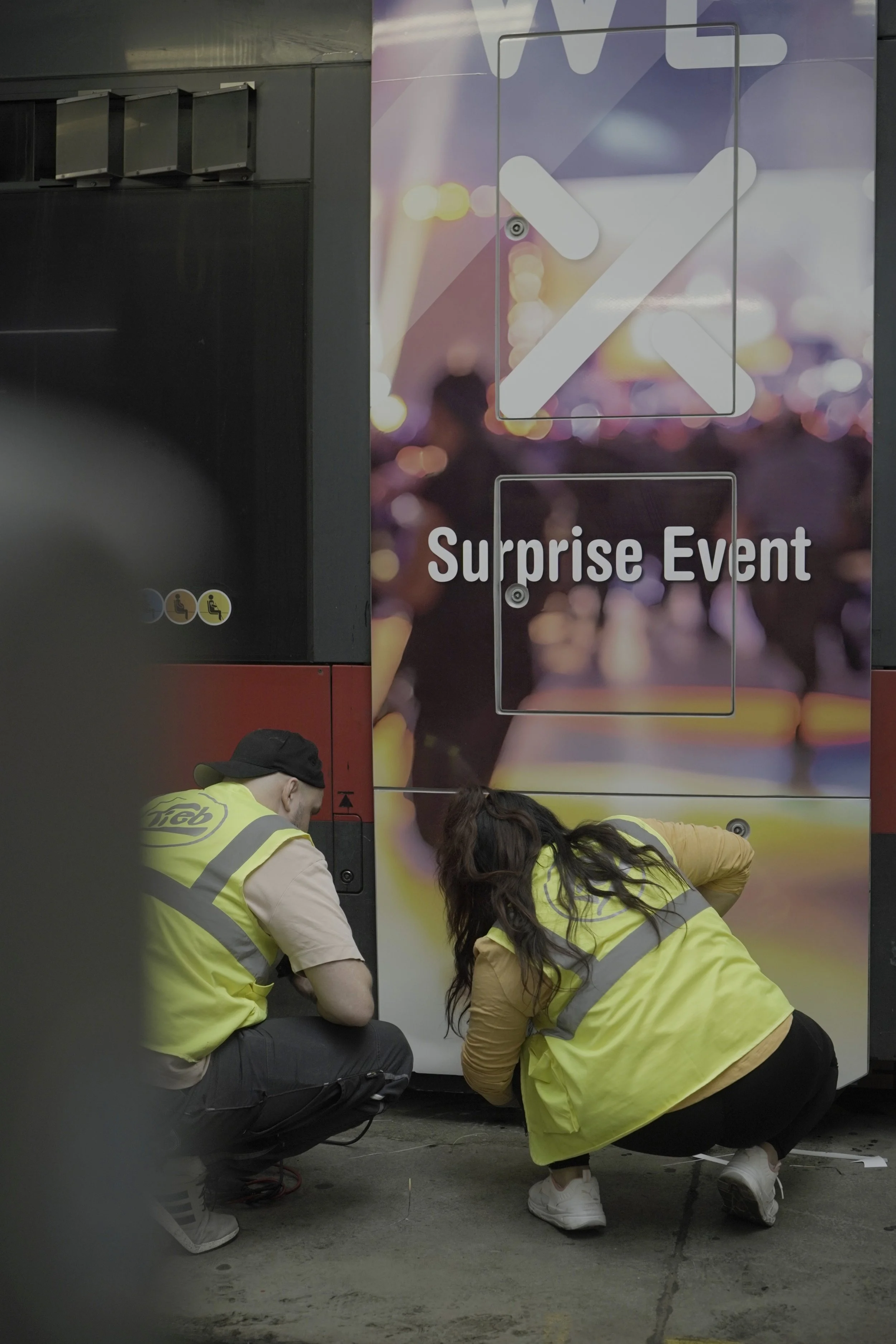 Two workers in yellow safety vests crouch down and work on a vending machine labeled 'Surprise Event' at a public space.