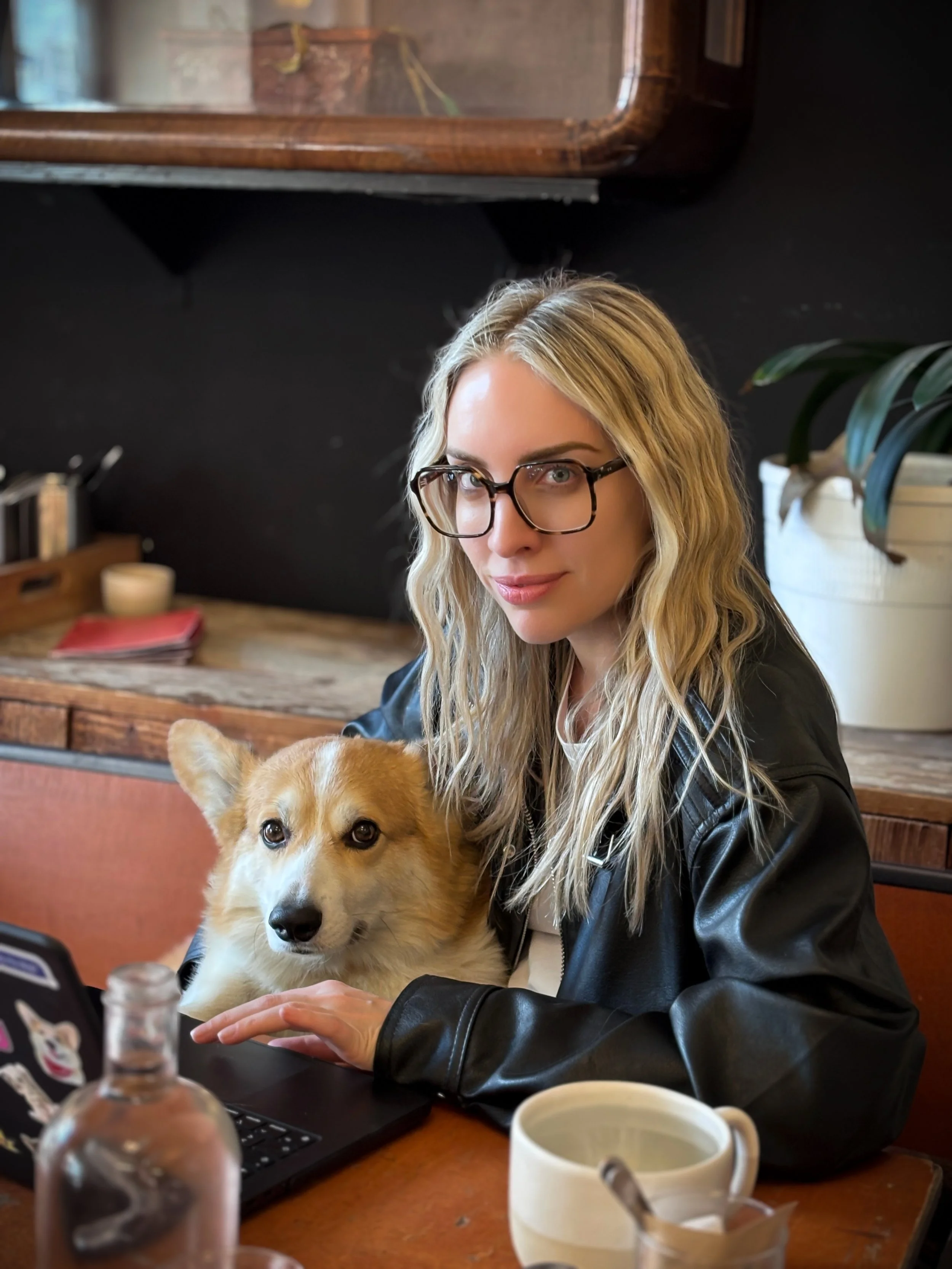 A woman with blonde wavy hair, black glasses, and a black leather jacket sitting at a table with a fluffy Corgi dog in a cozy indoor setting.