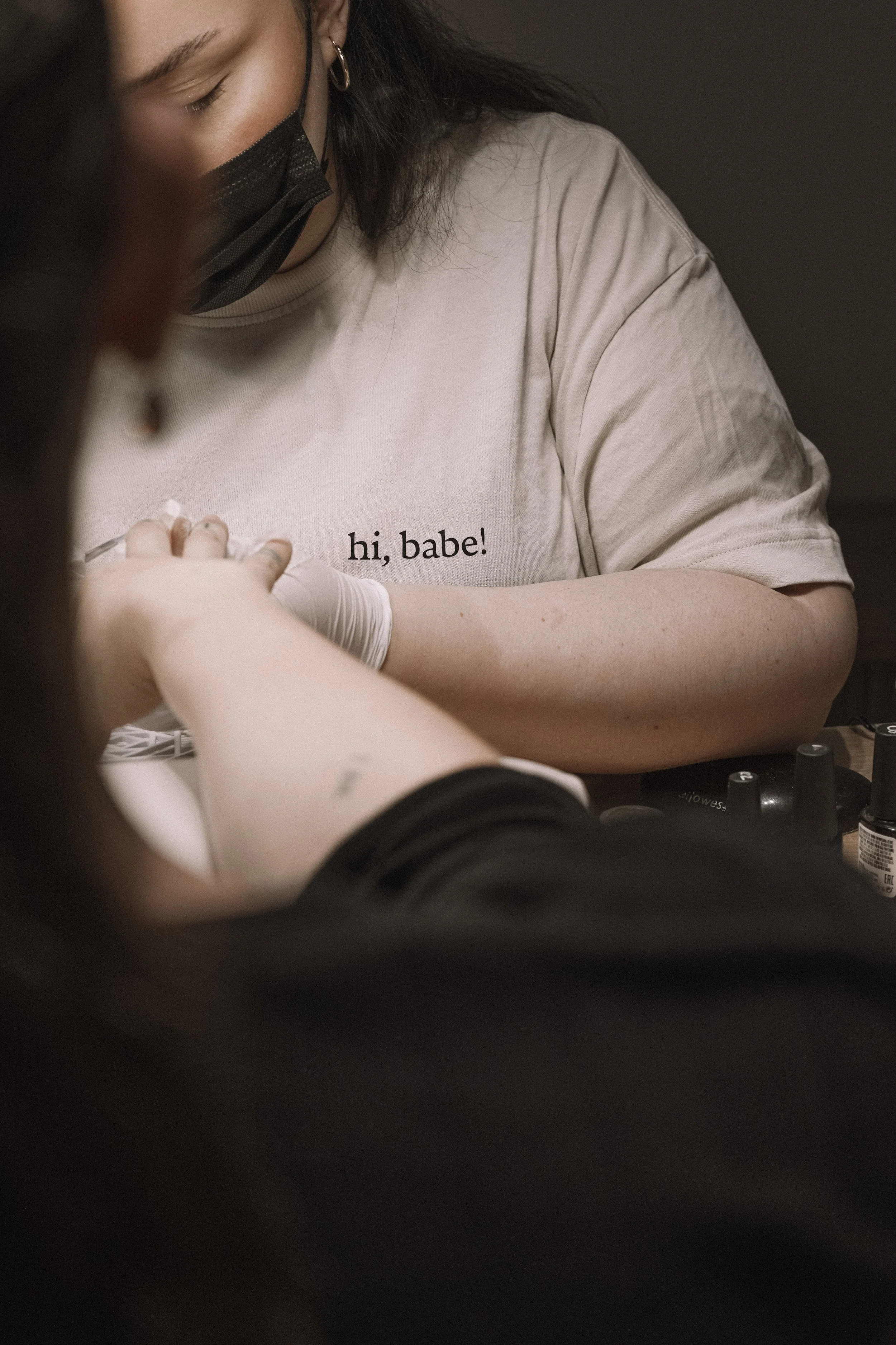 A person receiving a tattoo, wearing gloves, while another person with a beige shirt that says 'hi, babe!' observes. The person getting the tattoo is partially visible, and both are wearing face masks.