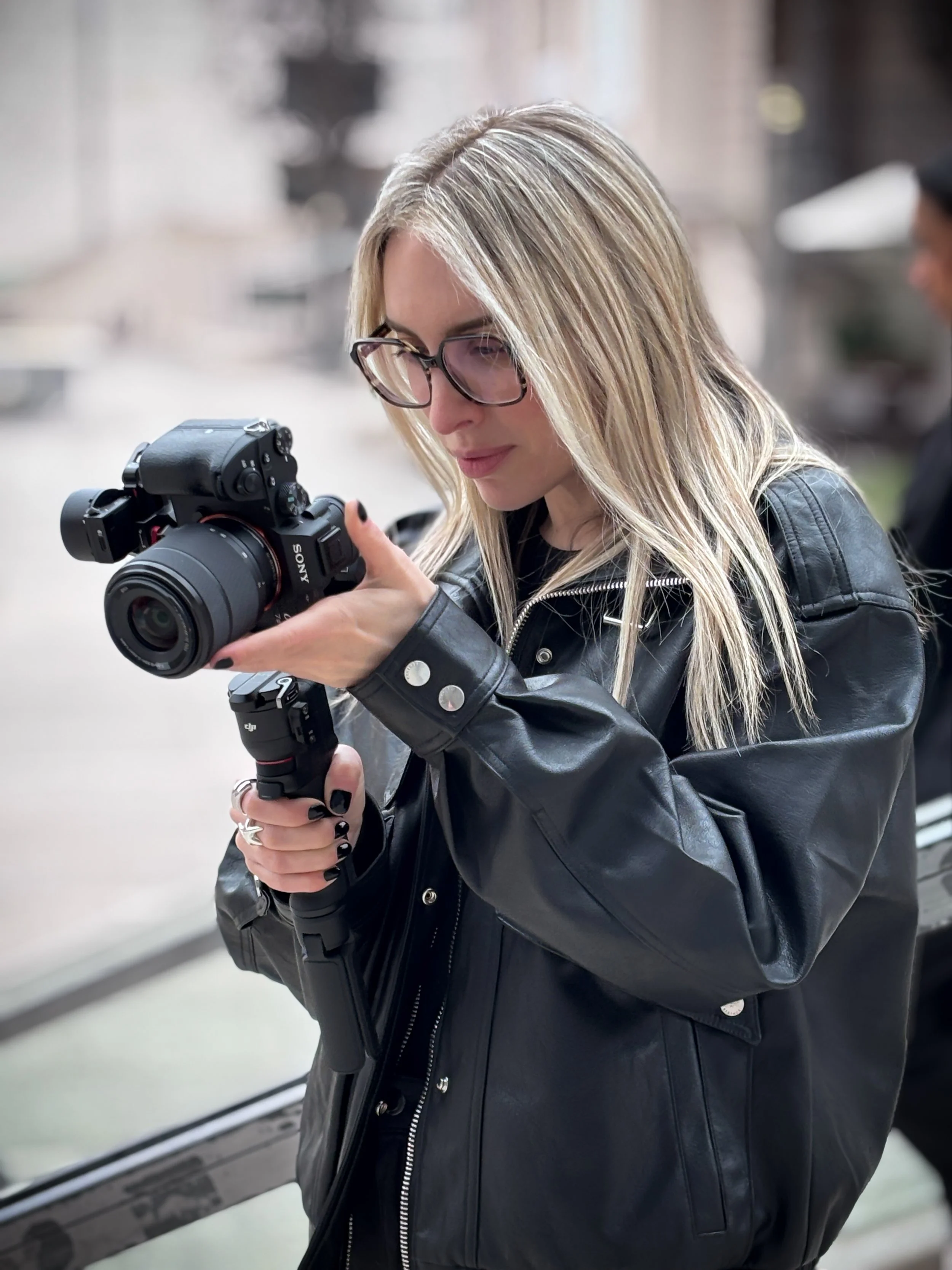 A woman with blonde hair, wearing glasses and a black leather jacket, is looking at a camera in her hands.