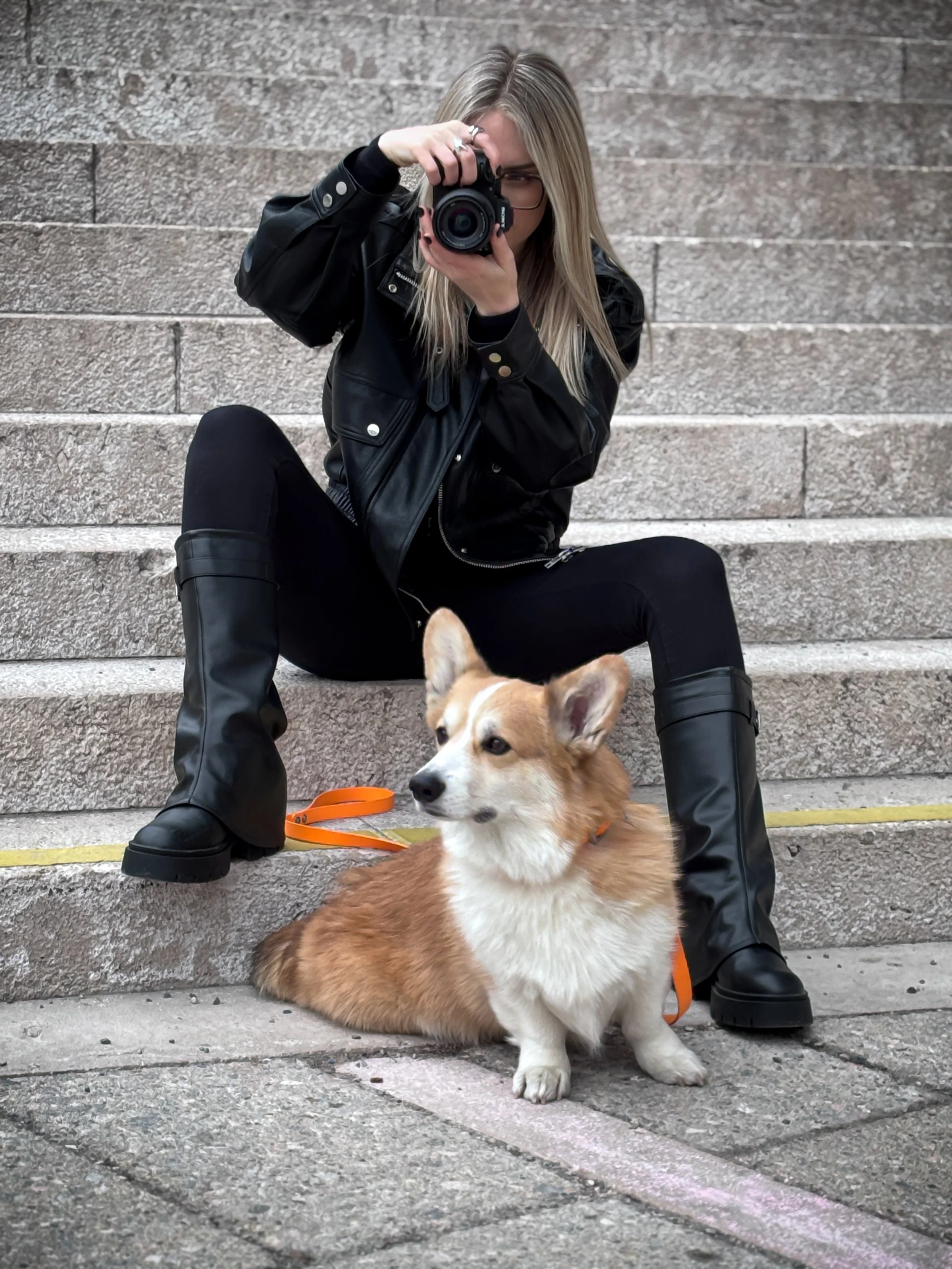 A woman in black leather jacket and black pants takes a photo with a camera while sitting on stone steps. Sitting beside her is a small, tan and white dog with pointed ears, a fluffy coat, and an orange leash.