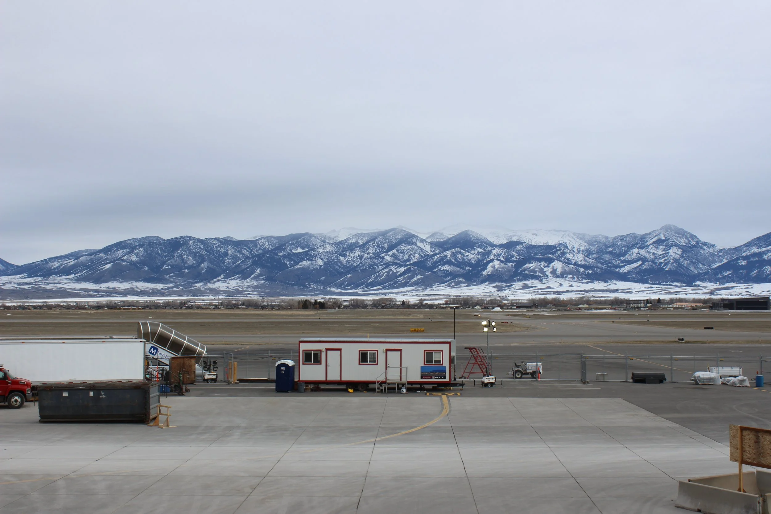 View of an airport tarmac with a portable building, vehicles, and equipment, with snow-covered mountains in the background under a cloudy sky.