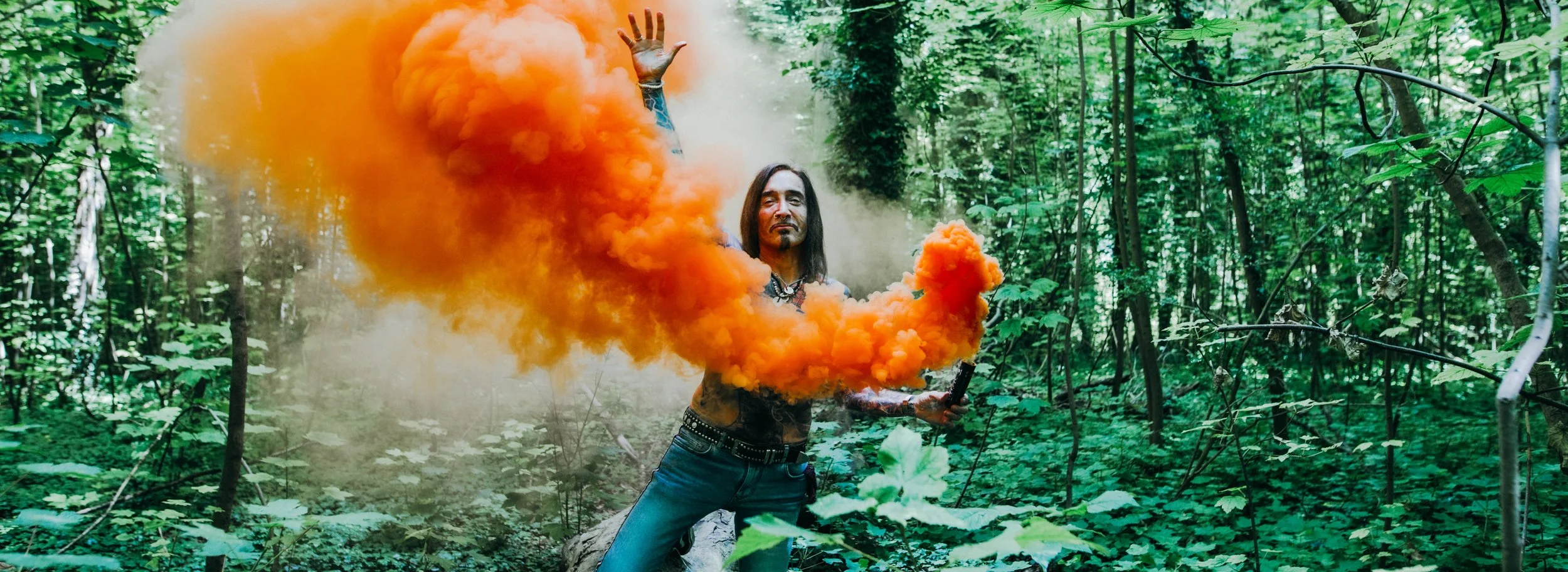 A man with long dark hair in a forest, holding a device, with orange smoke emitting from a smoke grenade.