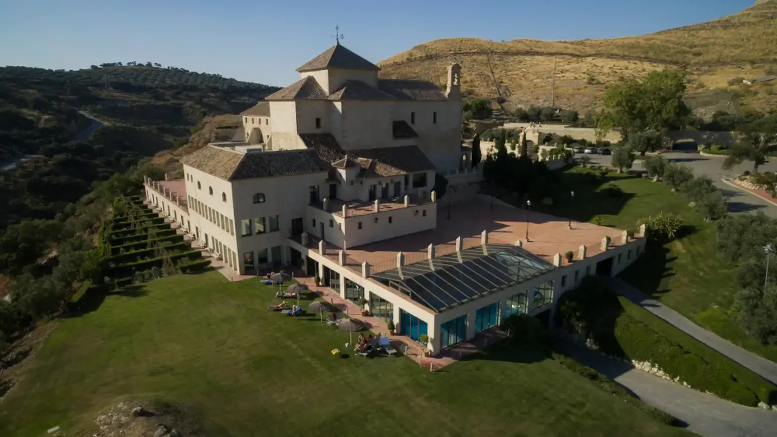 A large, castle-like building with a church on a hillside, surrounded by greenery and parking lot, under a clear blue sky.