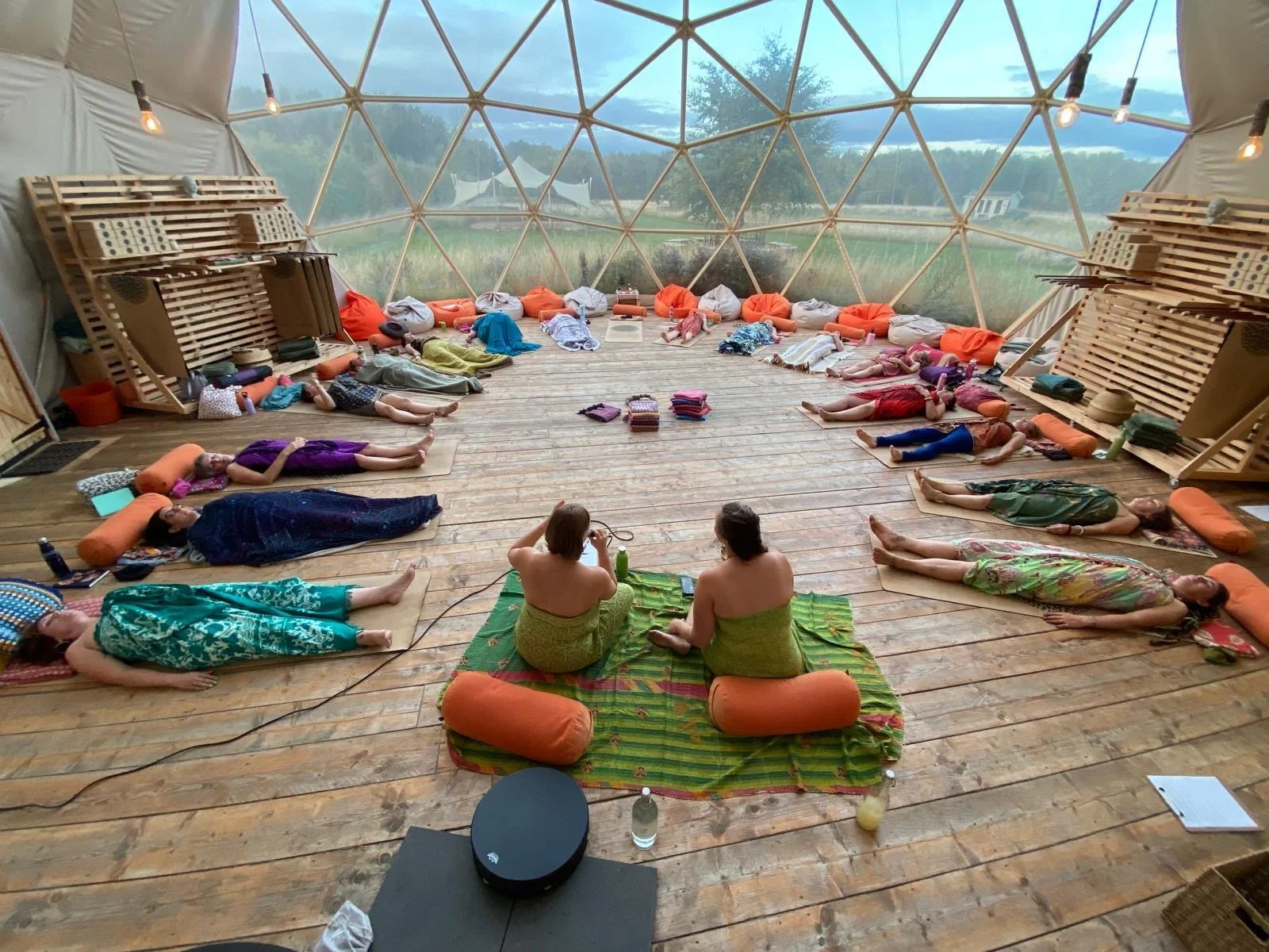 A group of people participating in a yoga or meditation class inside a geodesic dome structure with large windows, sitting or lying on mats with bolster pillows, overlooking a natural outdoor scenery.