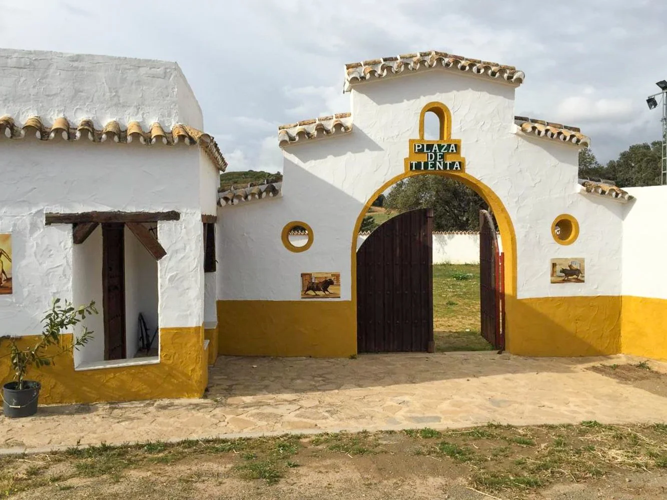 White colonial-style building with a yellow-trimmed archway entrance and a sign reading 'Plaza de Tienta'. The building features small circular windows and decorative paintings of horses on its walls, with a cobblestone pathway in front and a grassy area beyond.