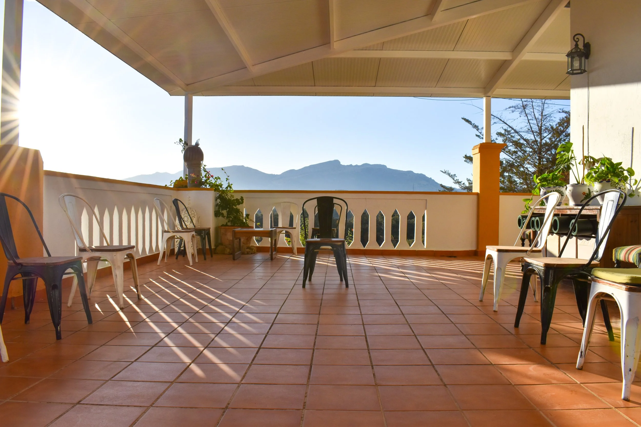 An outdoor terrace with terracotta tiles, surrounded by a white wall with decorative openings. Several chairs are arranged around the space, and potted green plants are on a table and on the floor. The background features mountain scenery and a clear sky, with sunlight casting shadows on the terrace.