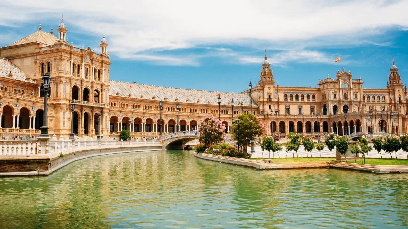Palace of the Parliament in Bucharest, Romania, with a water canal and pink flowering trees in the foreground under a partly cloudy sky.