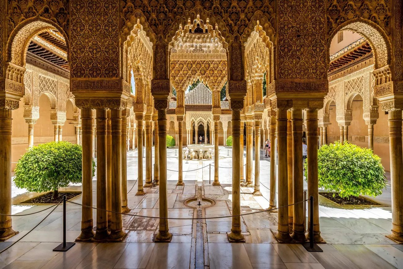 View of an ornate palace courtyard with intricately decorated arches, columns, and a fountain, featuring lush green plants and visitors in the background.