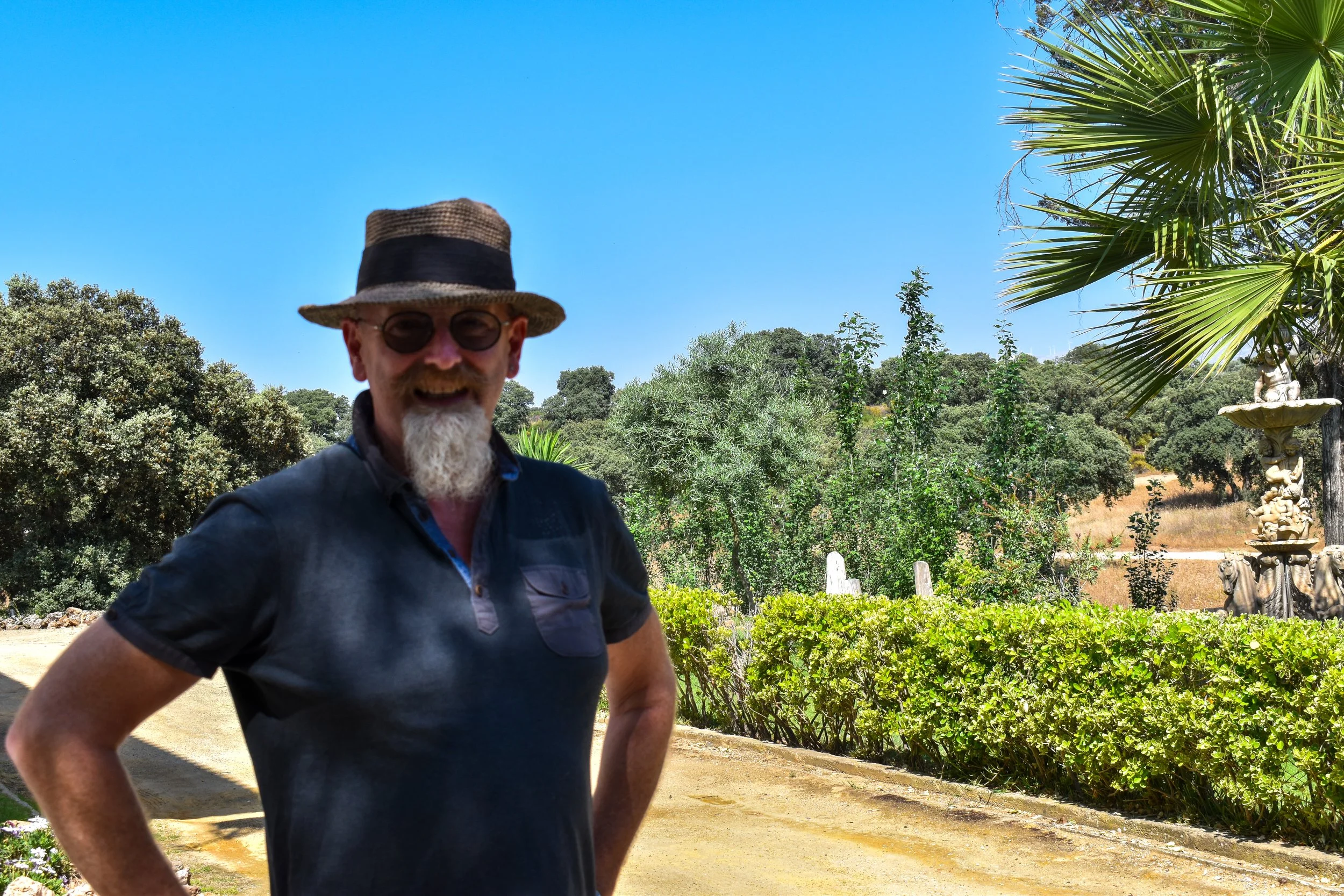 A man with a beard, wearing sunglasses, a straw hat, and a dark-colored polo shirt, standing outdoors with gardens, trees, and a fountain in the background under a clear blue sky.