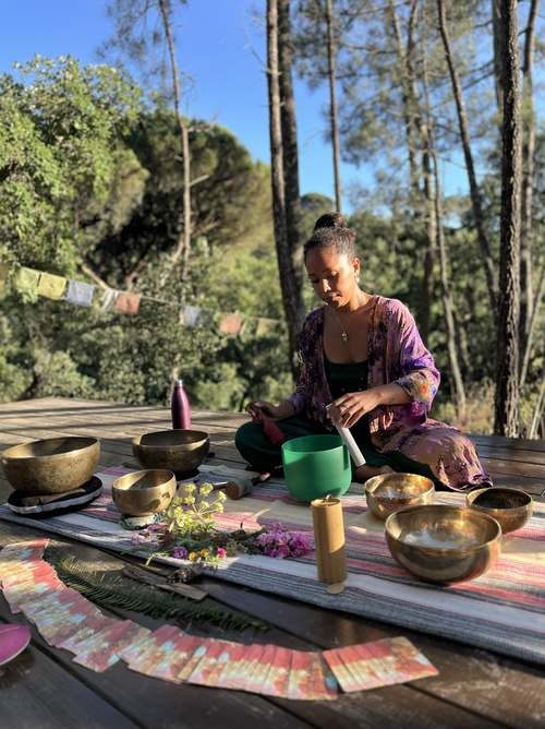 A woman sits cross-legged outdoors on a wooden platform, playing singing bowls with a mallet. She is surrounded by additional bowls, flowers, and a chart, with trees and a clear blue sky in the background.