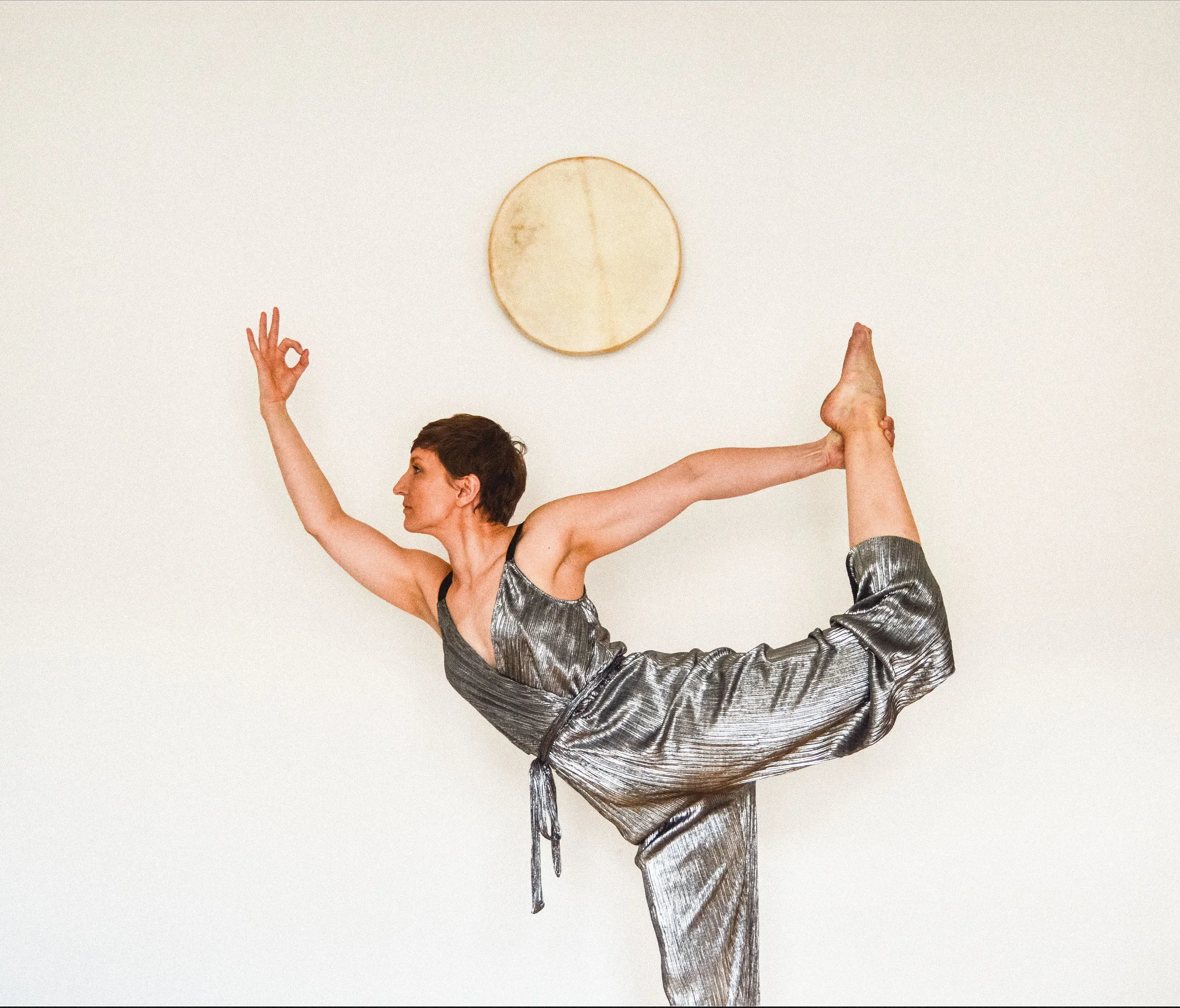 A woman practicing yoga, balancing on one leg while holding her foot behind her back with one hand and extending the other arm forward, in a pose called Lord of the Dance or Natarajasana, against a plain white wall with a round wooden wall decor.