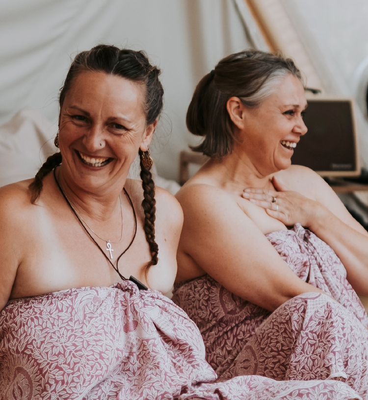 Two women sitting close together, wrapped in pink patterned towels, laughing and enjoying each other's company in a cozy room.