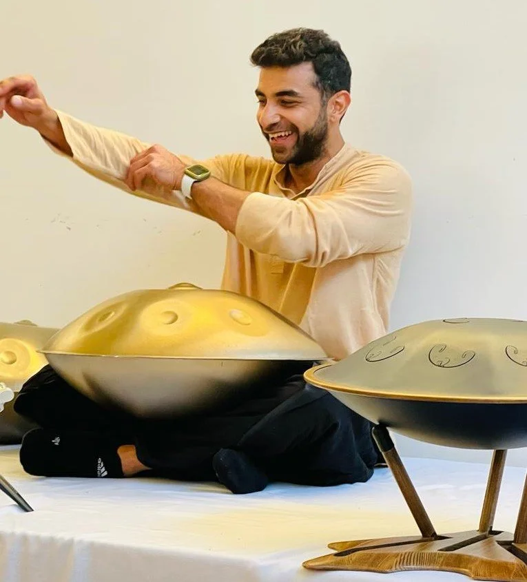 A man sitting cross-legged on a table, smiling, while playing a handpan with multiple other handpans around him.