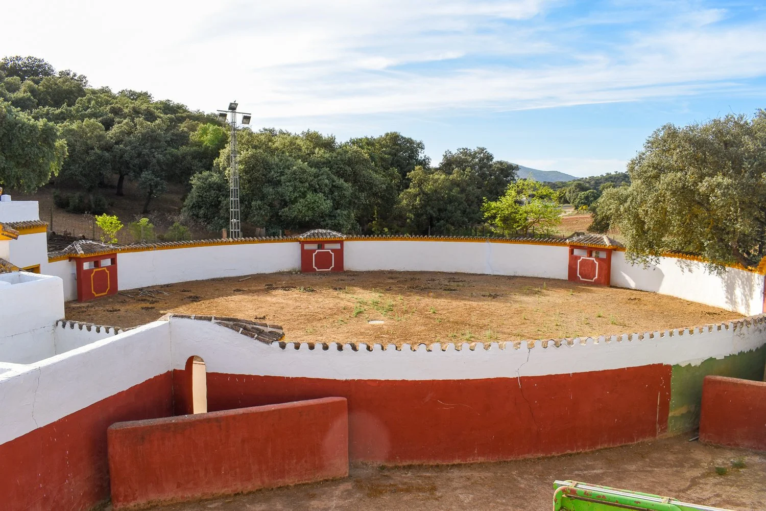 A bullring with white and red painted walls, surrounded by trees and hills in the background.