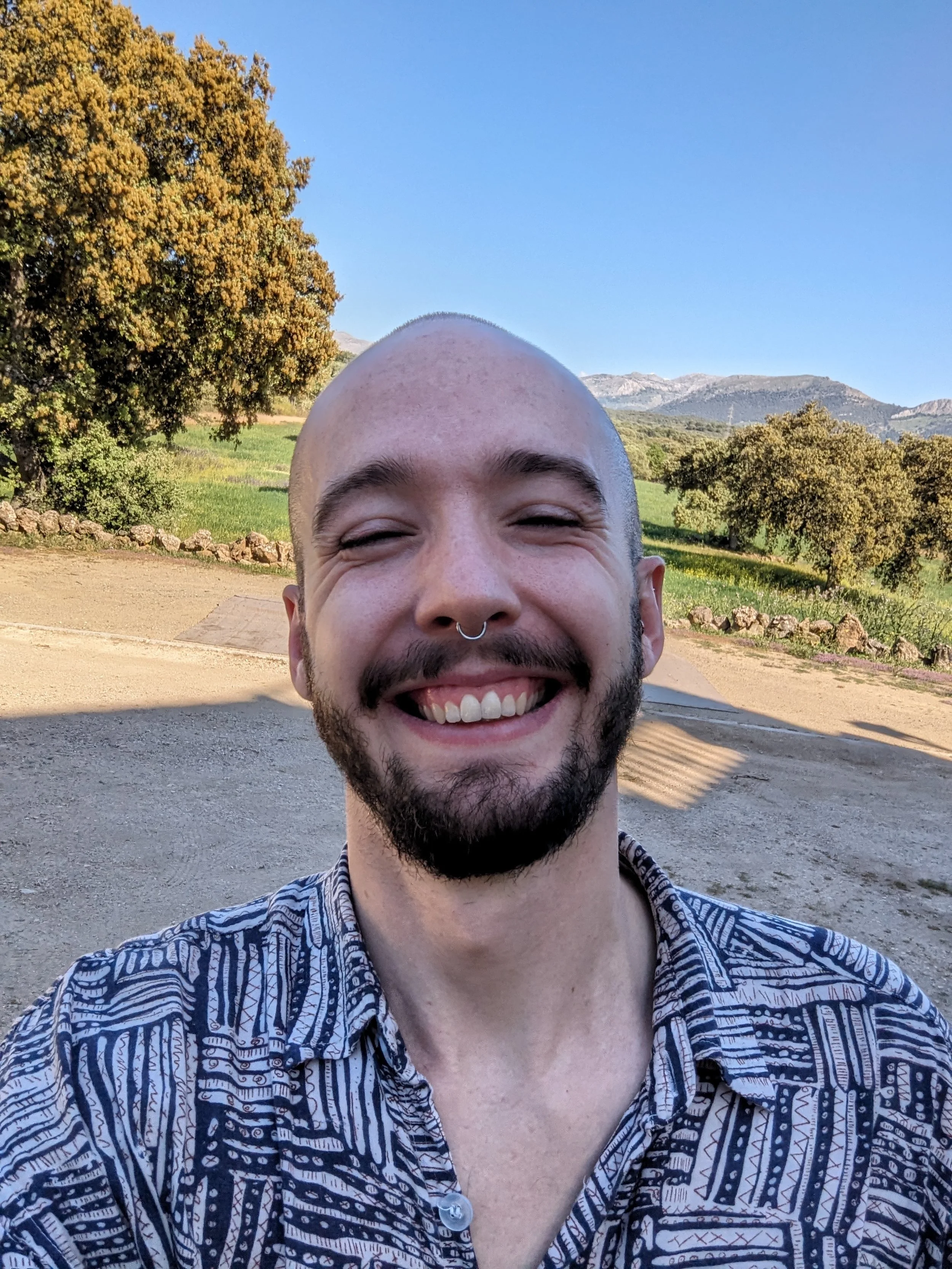 A smiling man with a beard and a septum piercing taking a selfie outdoors with green trees, mountains, and a clear blue sky in the background.