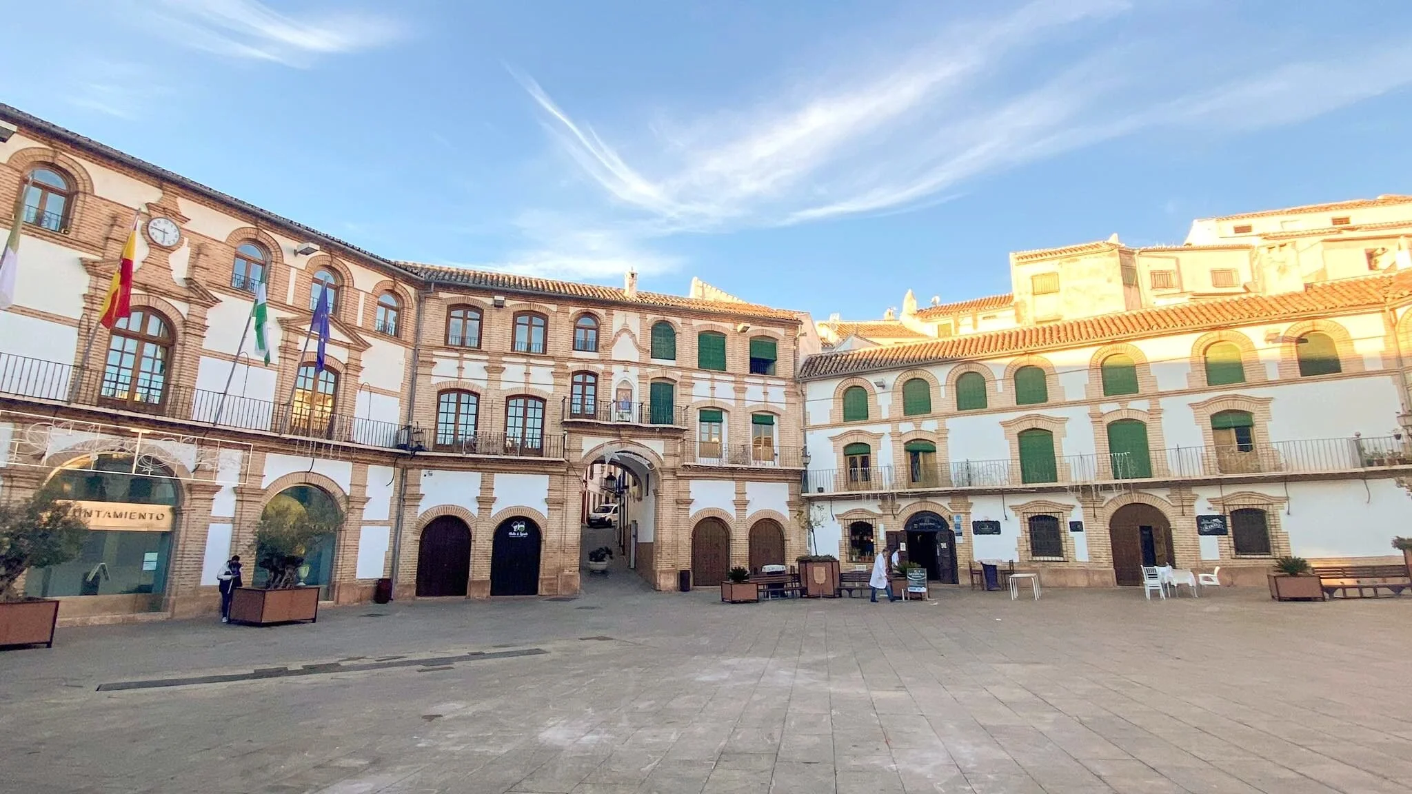 A town square with historic buildings, some with arched doorways and windows, flags hanging from the upper windows, and a few people walking or sitting. Clear blue sky overhead.