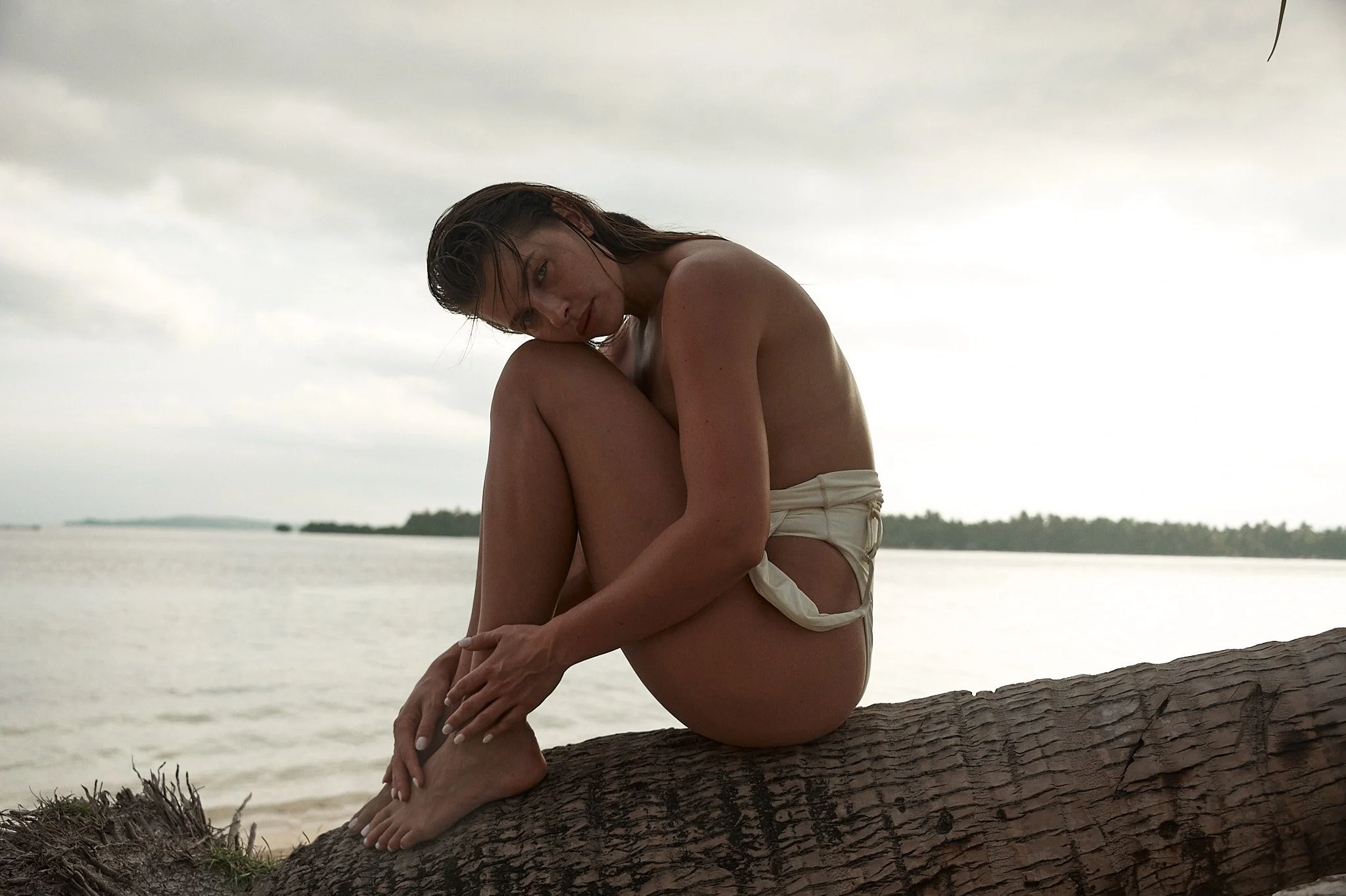 A woman sits on a large log at the beach, with her knees pulled up to her chest and her head resting on her knee, looking at the camera, under an overcast sky.