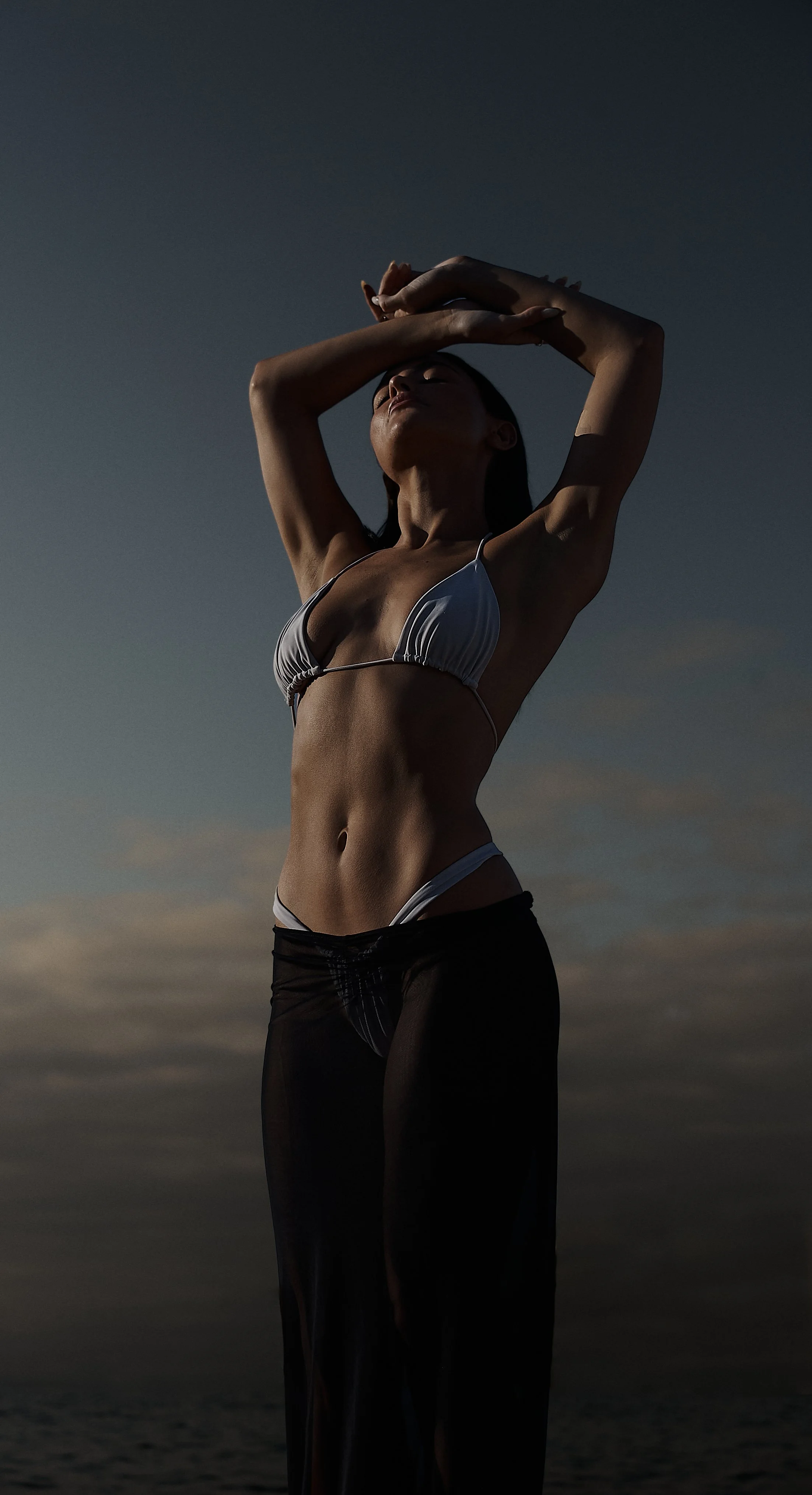 A woman in a bikini with dark sheer, mesh skirt standing outdoors at sunset or sunrise, raising her arms above her head with a cloudy sky in the background.
