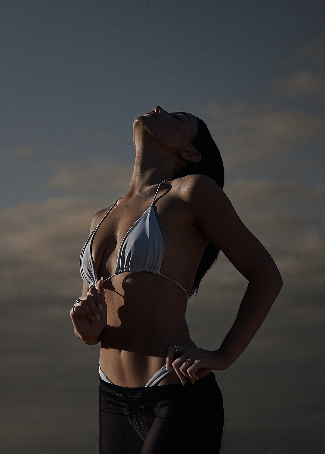 A woman in a white bikini stands outdoors against a cloudy sky, her face tilted upward with eyes closed, exhaling or basking in the sun.