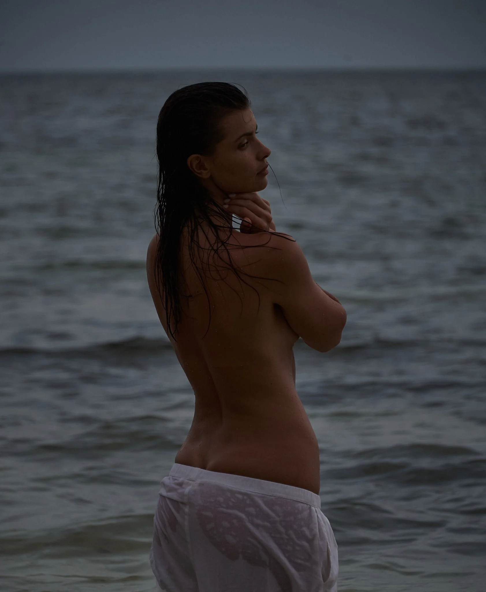A woman standing in the ocean, topless, with wet hair, wearing white shorts, looking to the side at dusk or dawn.