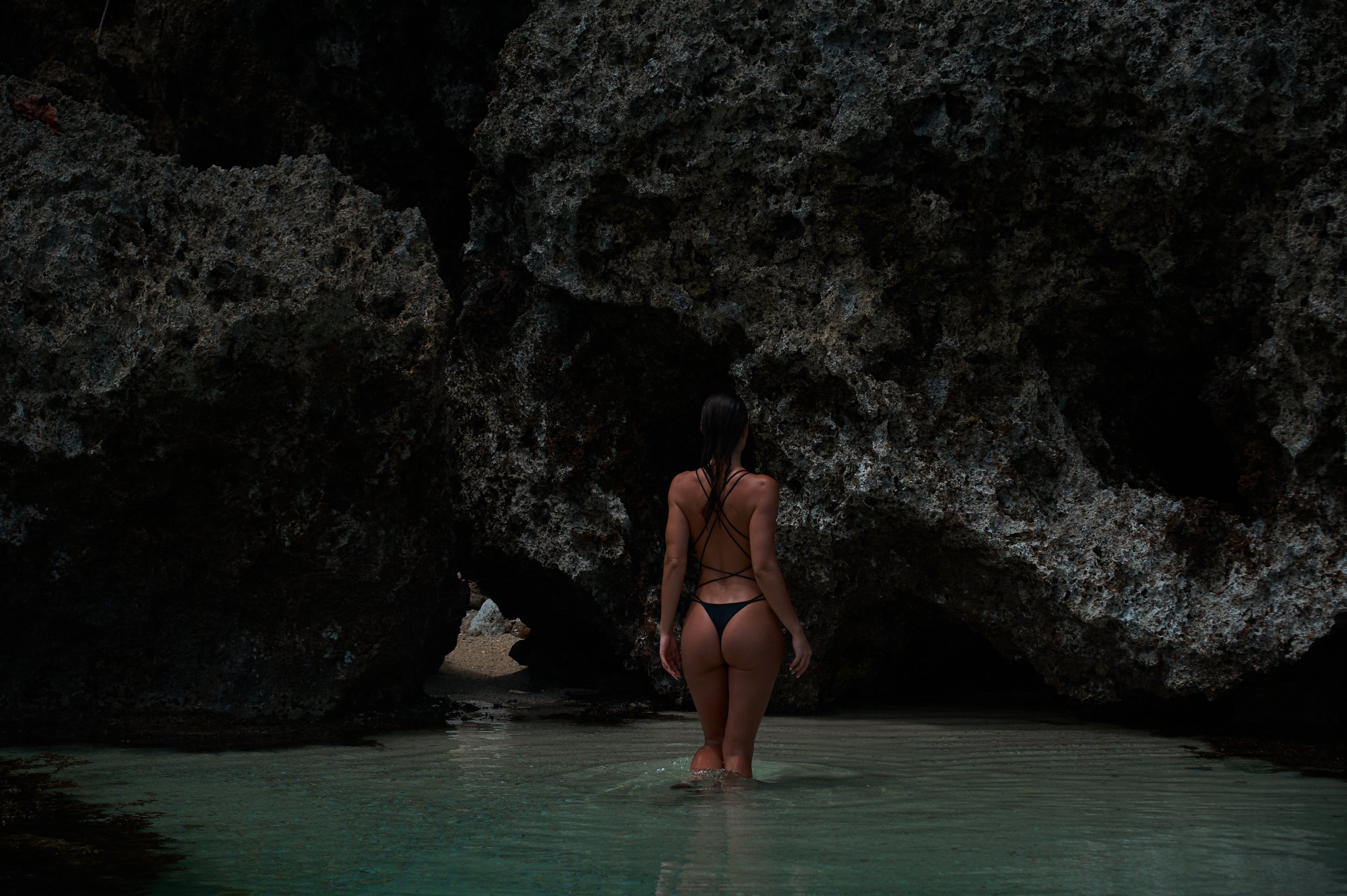 Woman in a black bikini standing in water near large rocky formations at the beach.