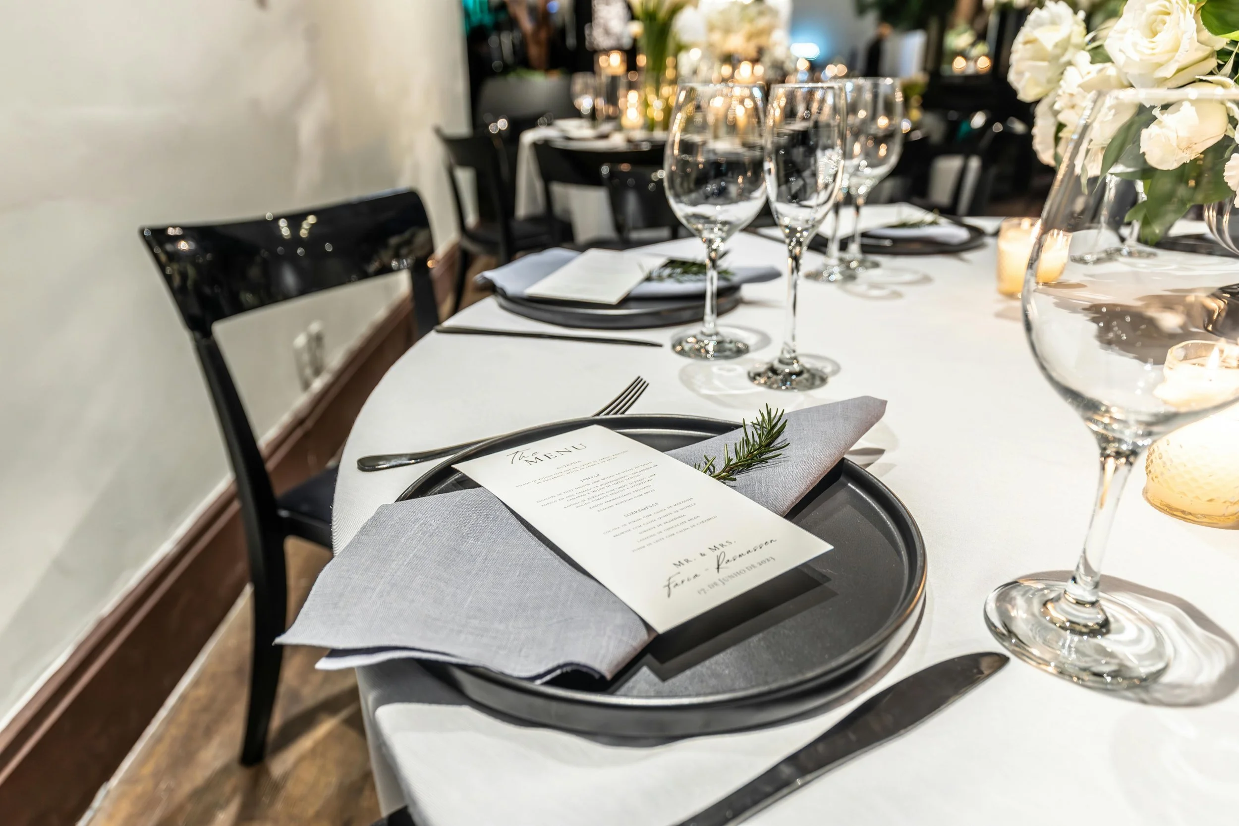 Elegant table setting with black plates, silverware, wine glasses, and a menu, decorated with a sprig of rosemary, for a formal event or dinner. White tablecloth and floral centerpiece with candles.