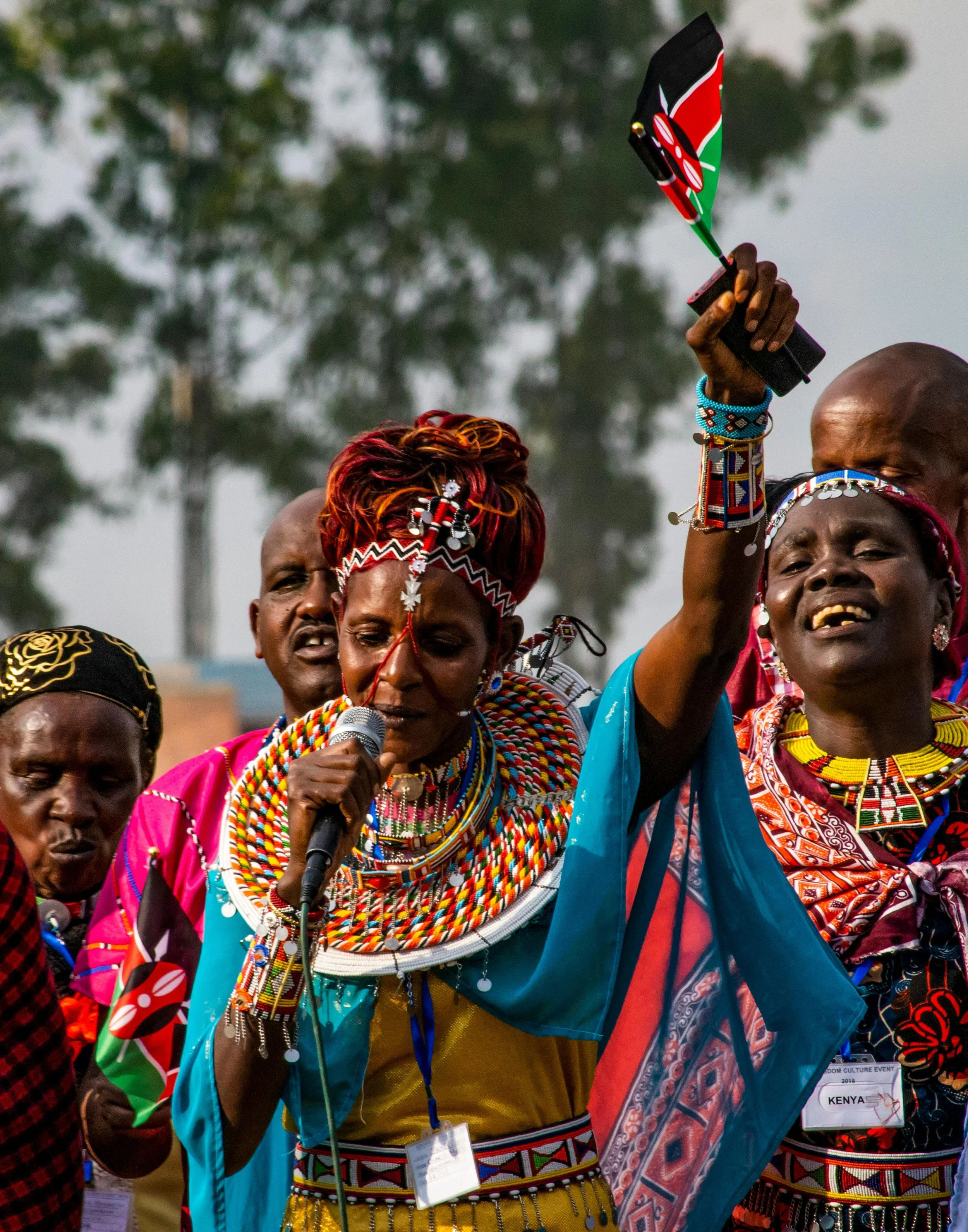 A group of Kenyan people dressed in traditional colorful Maasai attire, with one woman holding a microphone and a small Kenyan flag, celebrating at an outdoor event.