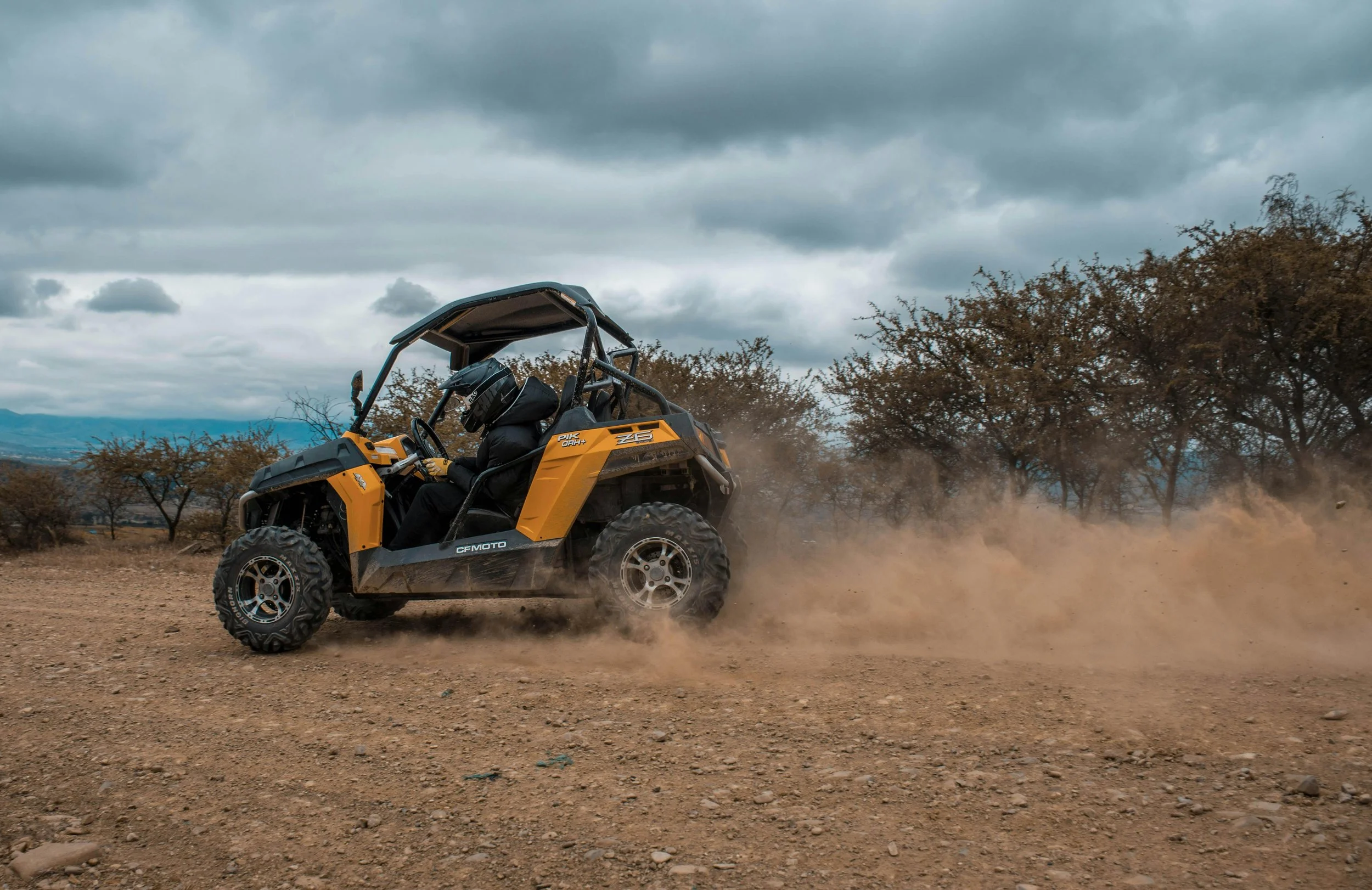 An off-road vehicle driving across rocky dirt terrain with dust trailing behind, under a cloudy sky.