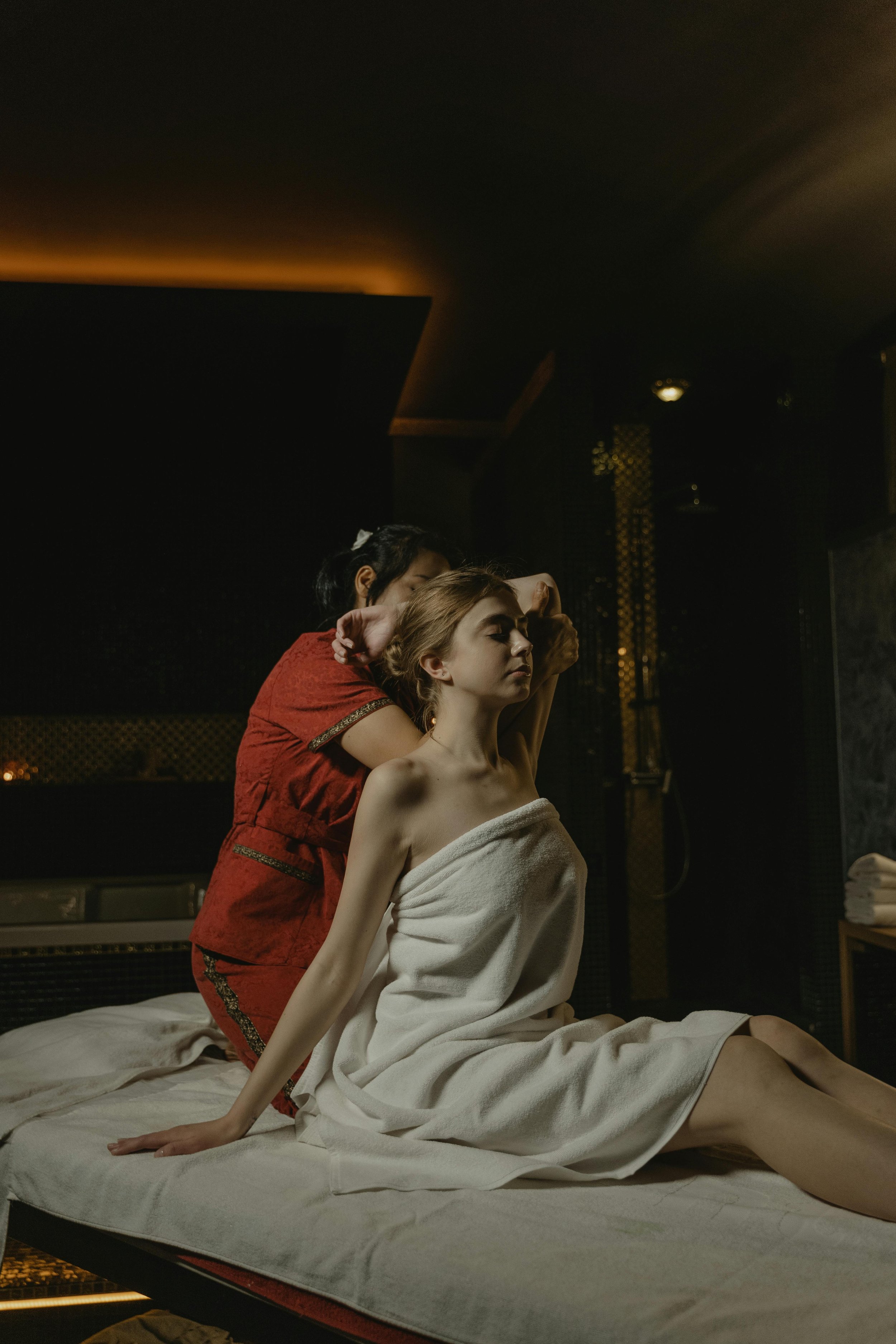 A woman sitting on a bed wrapped in a towel receives a massage from a woman in a red uniform in a dimly lit spa room.