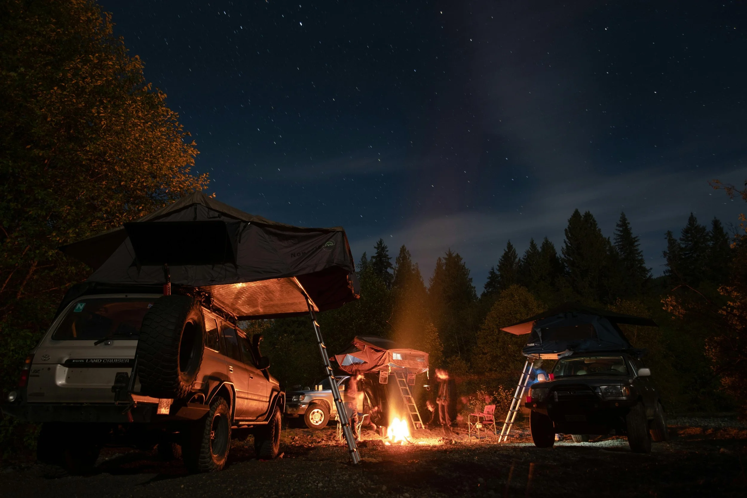 Two off-road vehicles with rooftop tents parked around a campfire at night in a wooded area, with a star-filled sky overhead.
