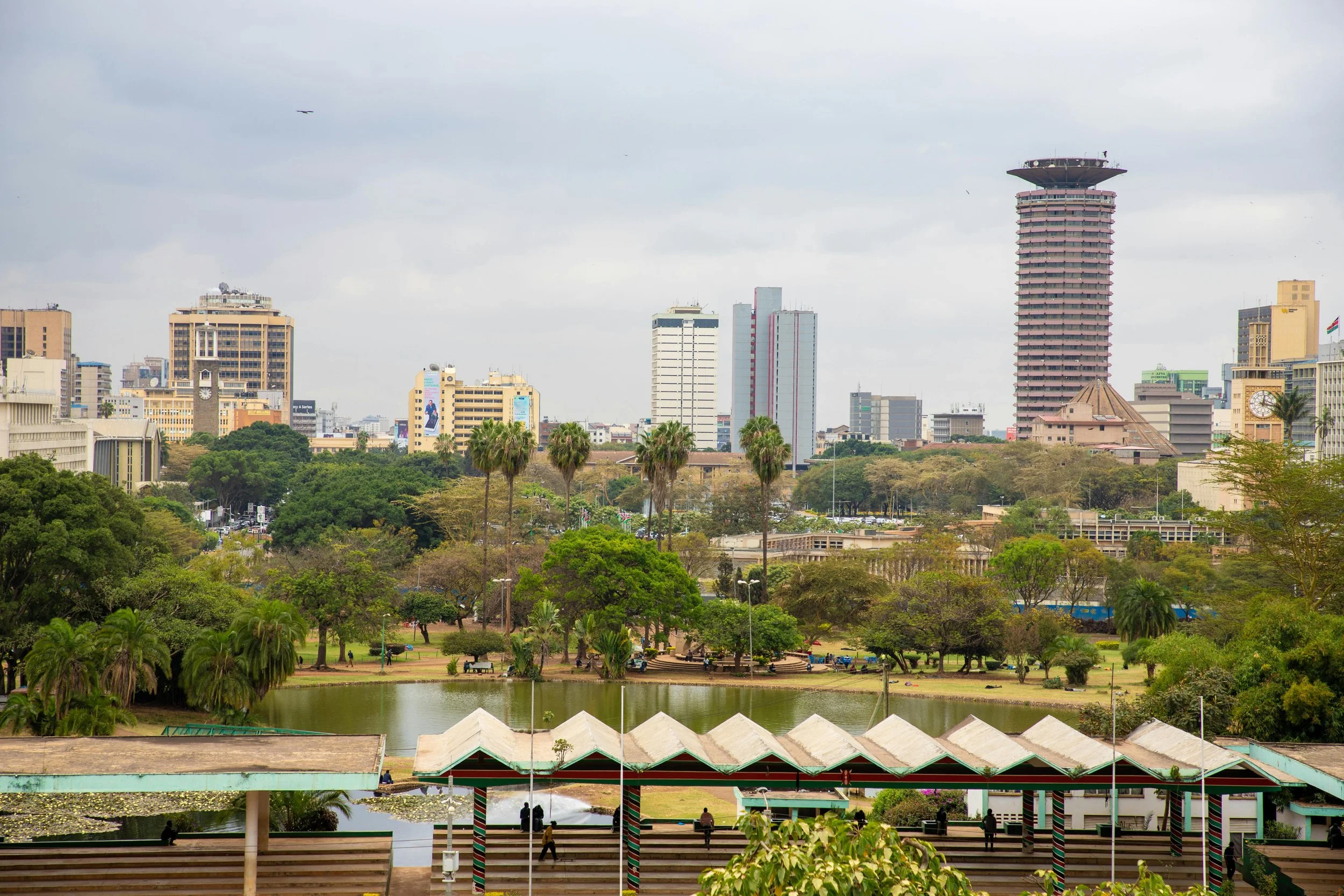 Cityscape with tall buildings, green trees, a small pond, and a pavilion with a zigzag roof in a park setting.