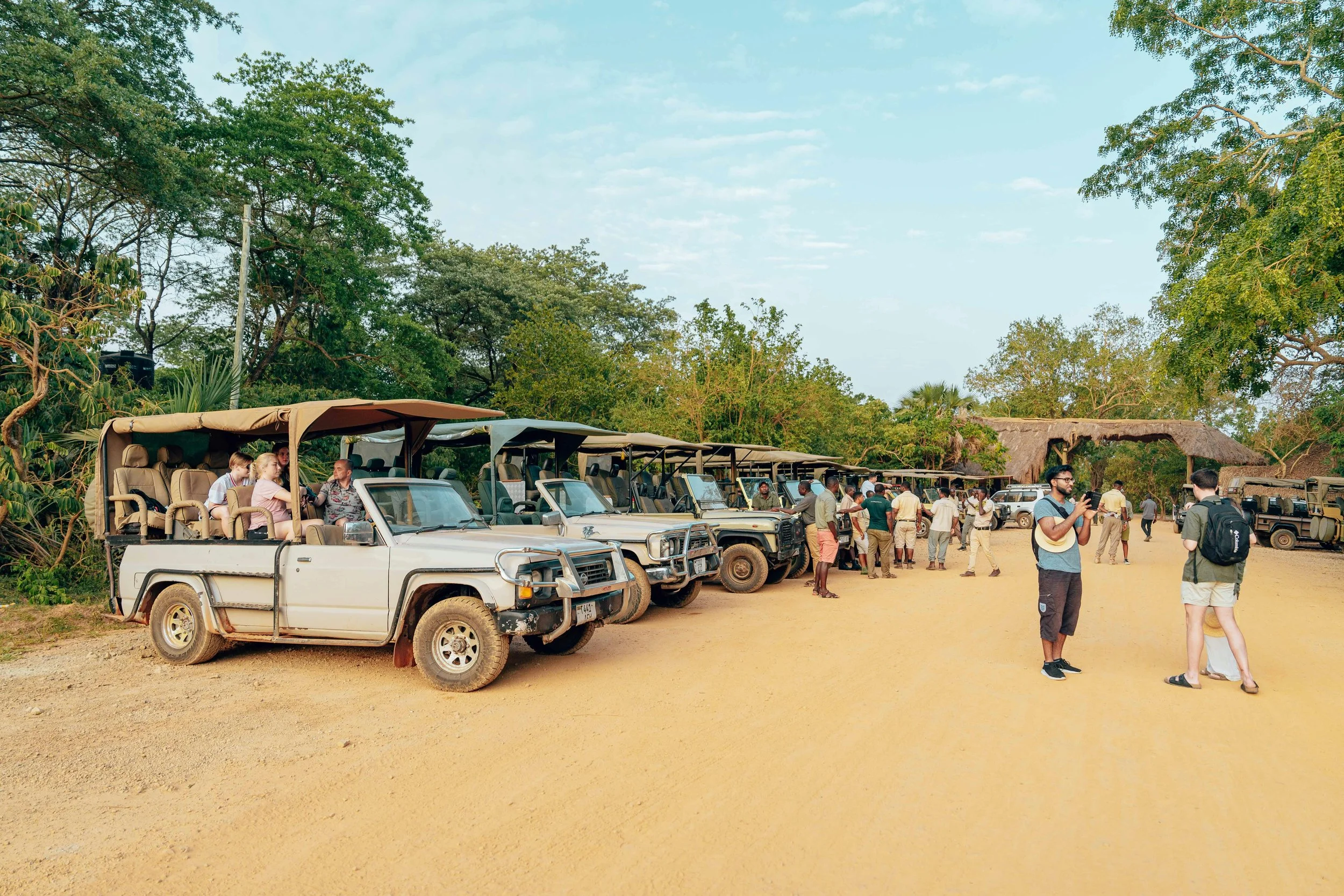 Three safari vehicles driving along a dirt road in a savanna landscape with trees, with a hot air balloon floating in the sky above.