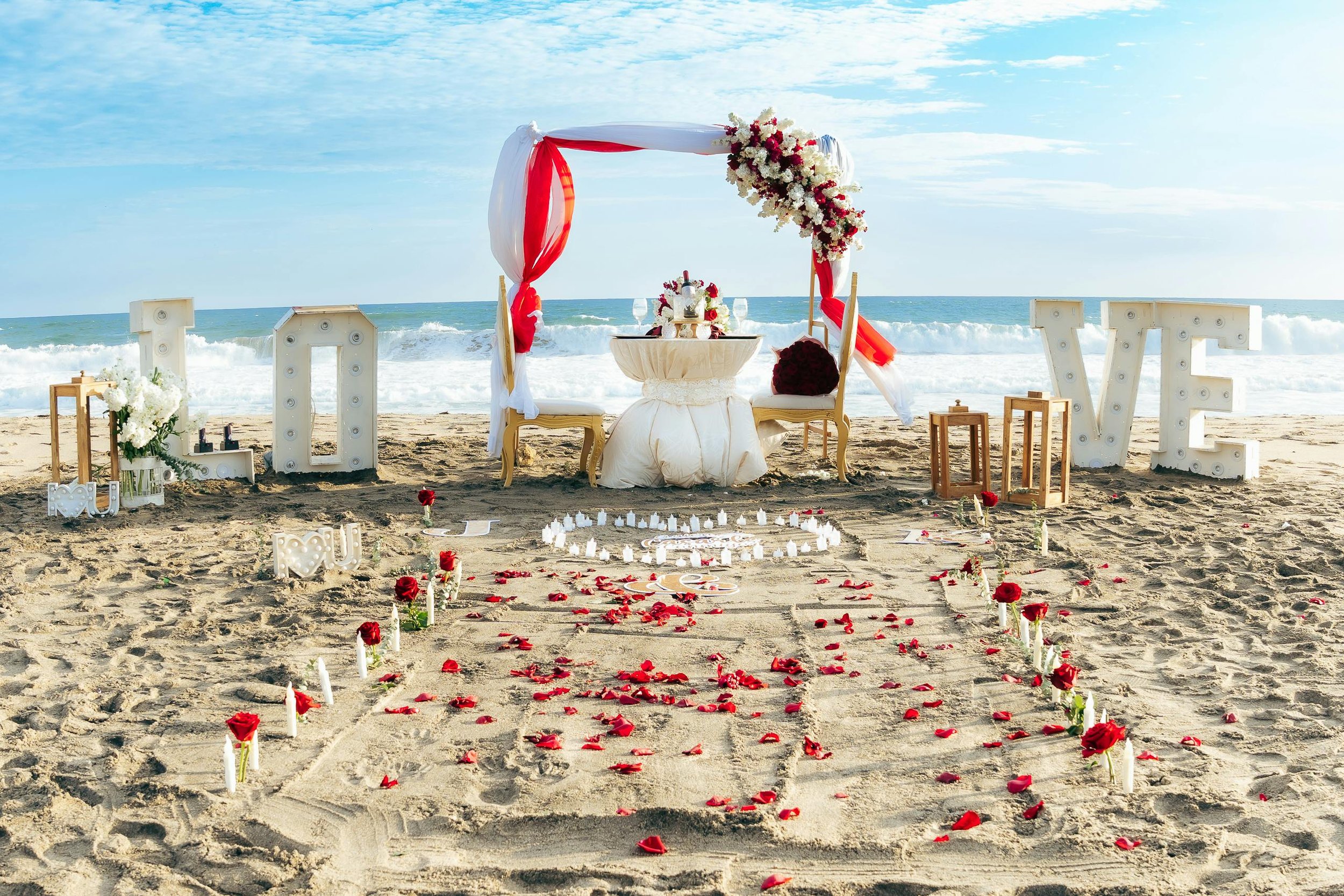 Beach wedding setup with an arch decorated with flowers, a table, chairs, large illuminated letters spelling love, candles, and rose petals on the sand facing the ocean.