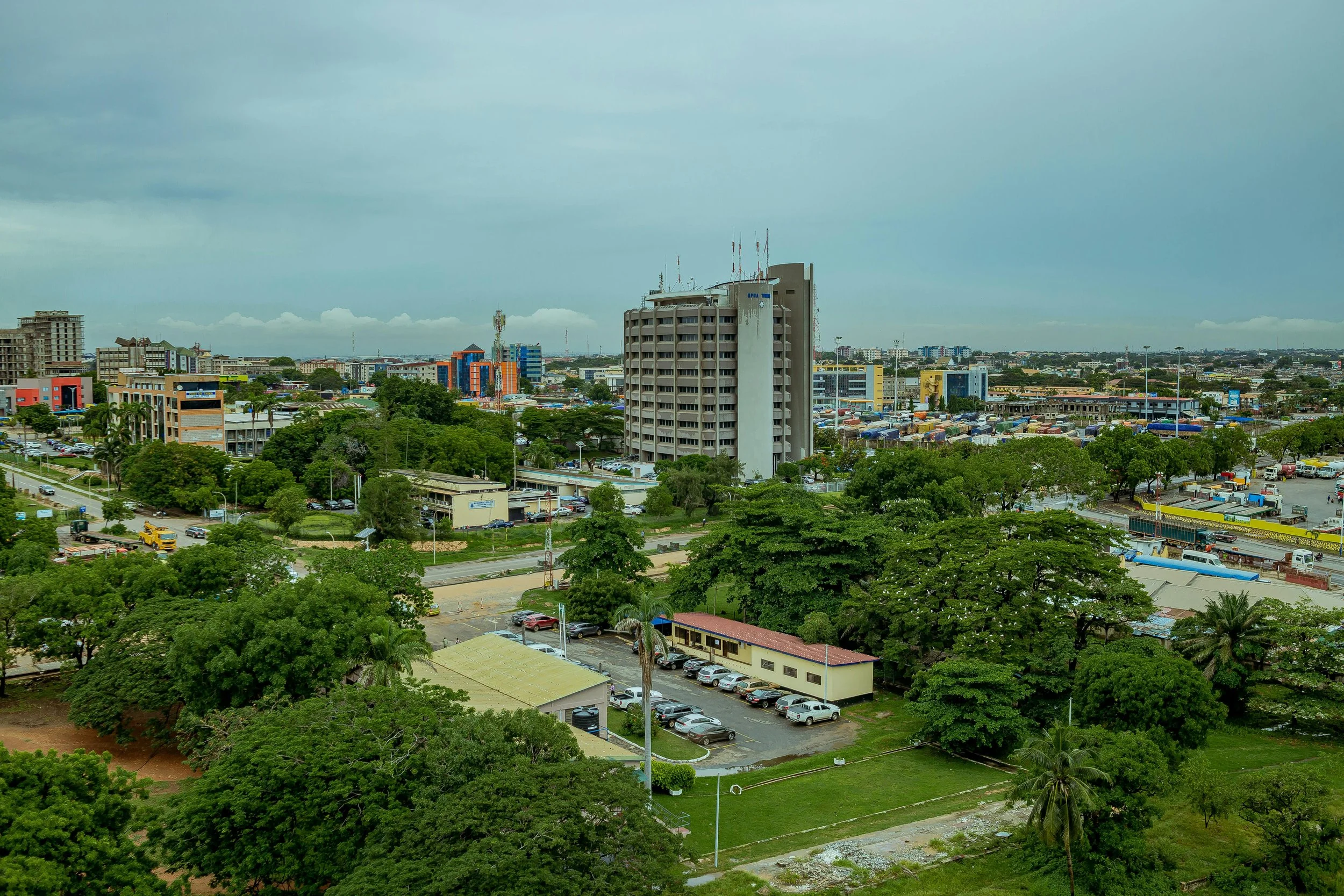 A cityscape with multi-story buildings, trees, parking lots, and roads under a cloudy sky.