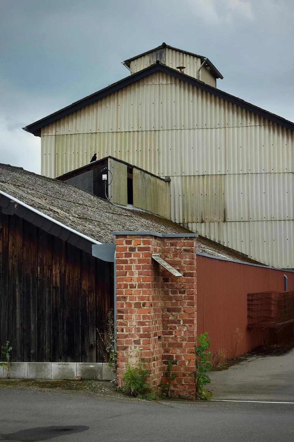 A rustic brick pillar on the street corner next to a black wooden fence and red wall, with a weathered rooftop, a bird perched on the building, and a large cream-colored industrial building in the background under a cloudy sky.