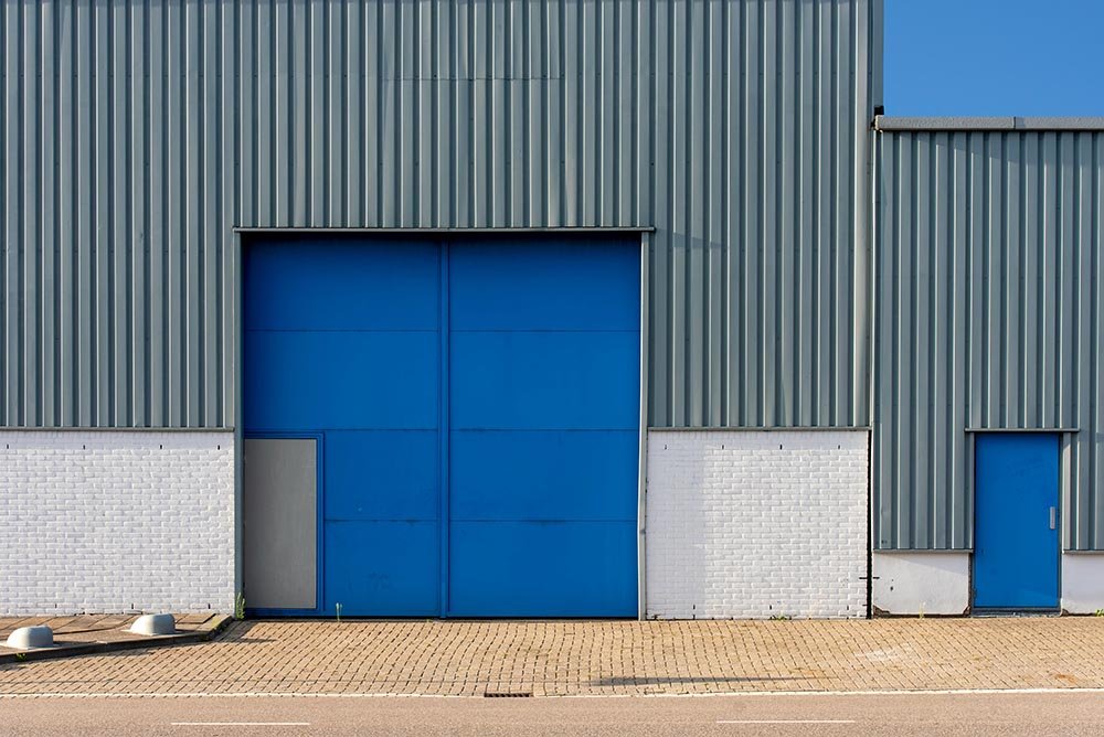 Industrial building with blue metal facade and large blue sliding door, small gray door, white brick wall, and paved sidewalk.