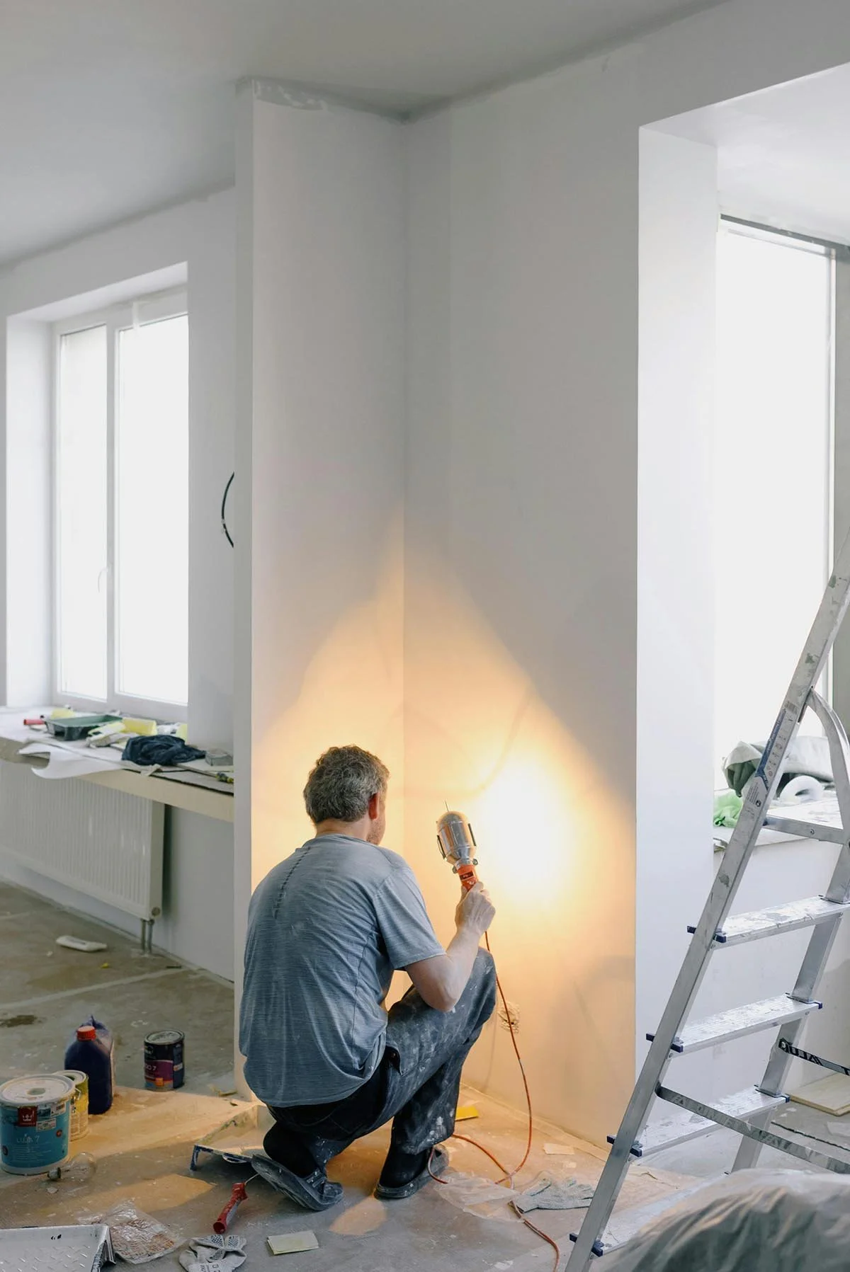 A person kneeling on the floor with a tool in hand, working on an interior wall in a room with large windows, ladders, and construction supplies scattered around.