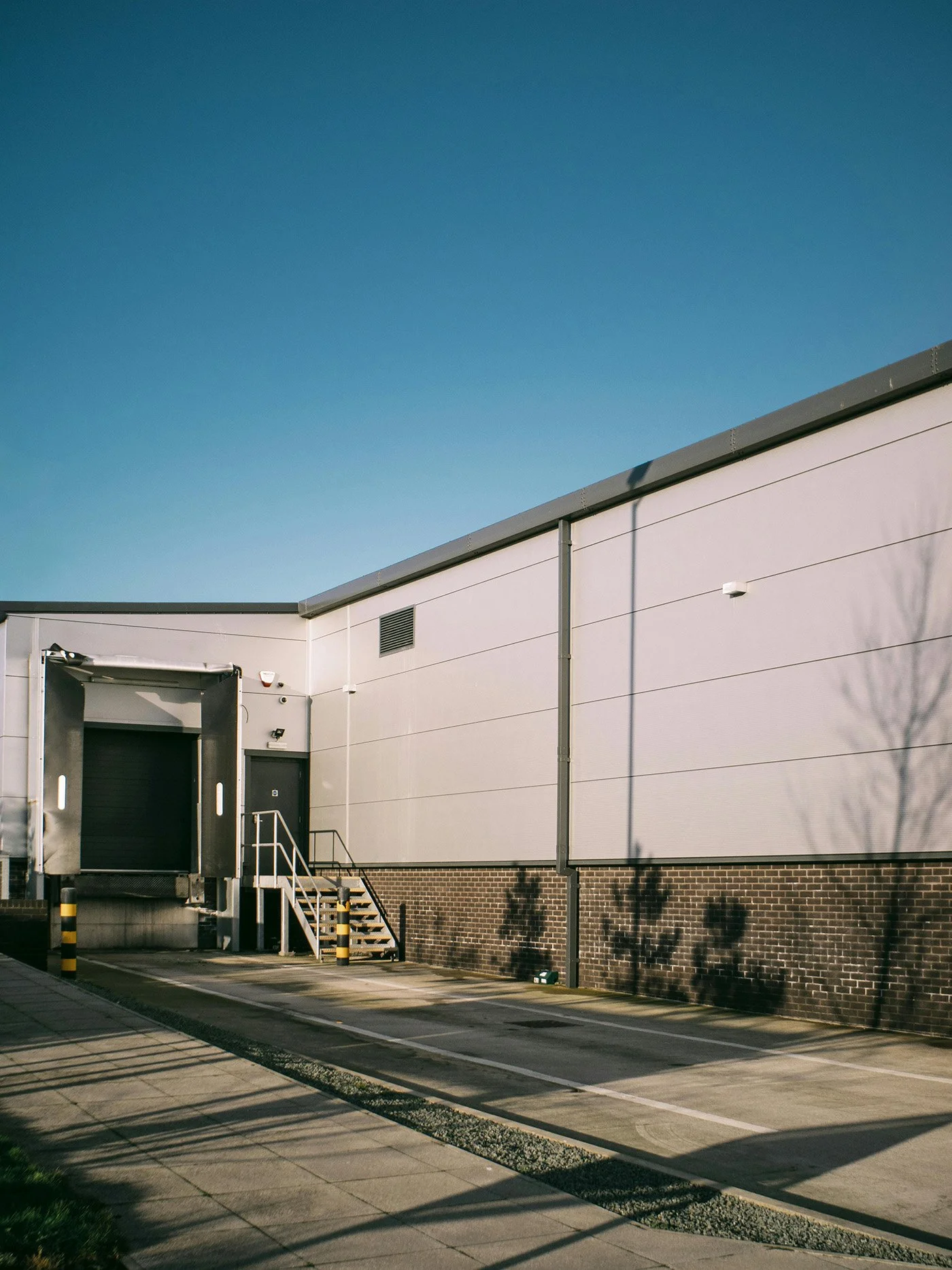 Exterior of a warehouse building with a loading dock, stairs, and parking lot shadows on the ground, under a clear blue sky.