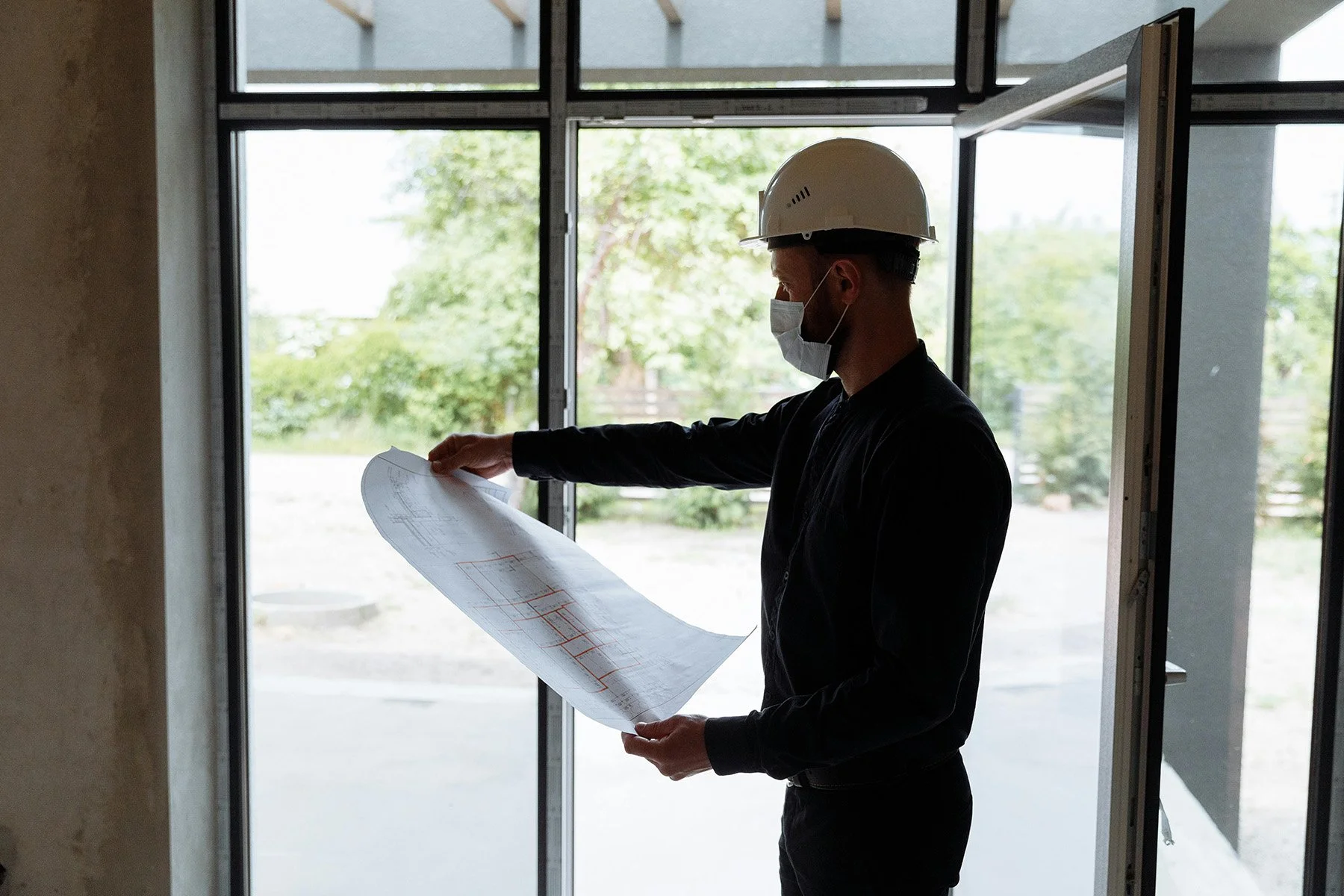 A construction worker wearing a white hard hat and face mask reviews blueprints inside a building under construction, with large windows showing trees outside.