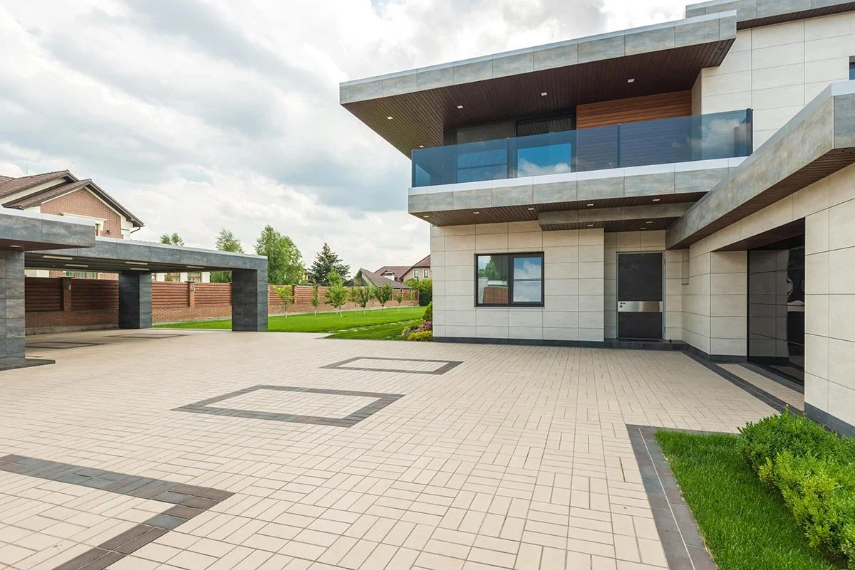 Modern house with a tiled outdoor patio area, including a covered parking space, green lawn, and landscaping. The house features large windows, balconies with glass railings, and contemporary architectural design.