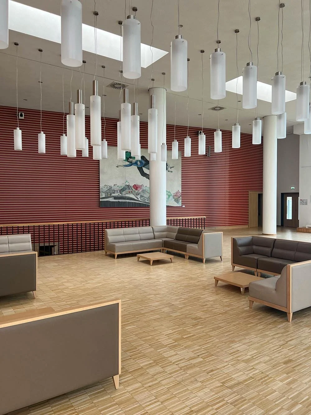 Modern indoor lounge area with neutral-colored sofas and small coffee tables, a red slatted wall, a large wall art, and hanging cylindrical pendant lights.