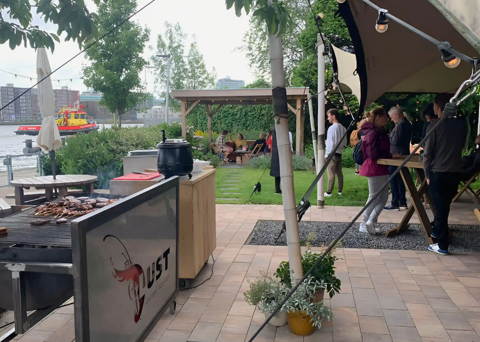 Mensen wachten in een rij bij een buitenterras met een bier- en grillstation, voorzien van een afdak en tuinmeubels, met een rivier en een rivierboot op de achtergrond.