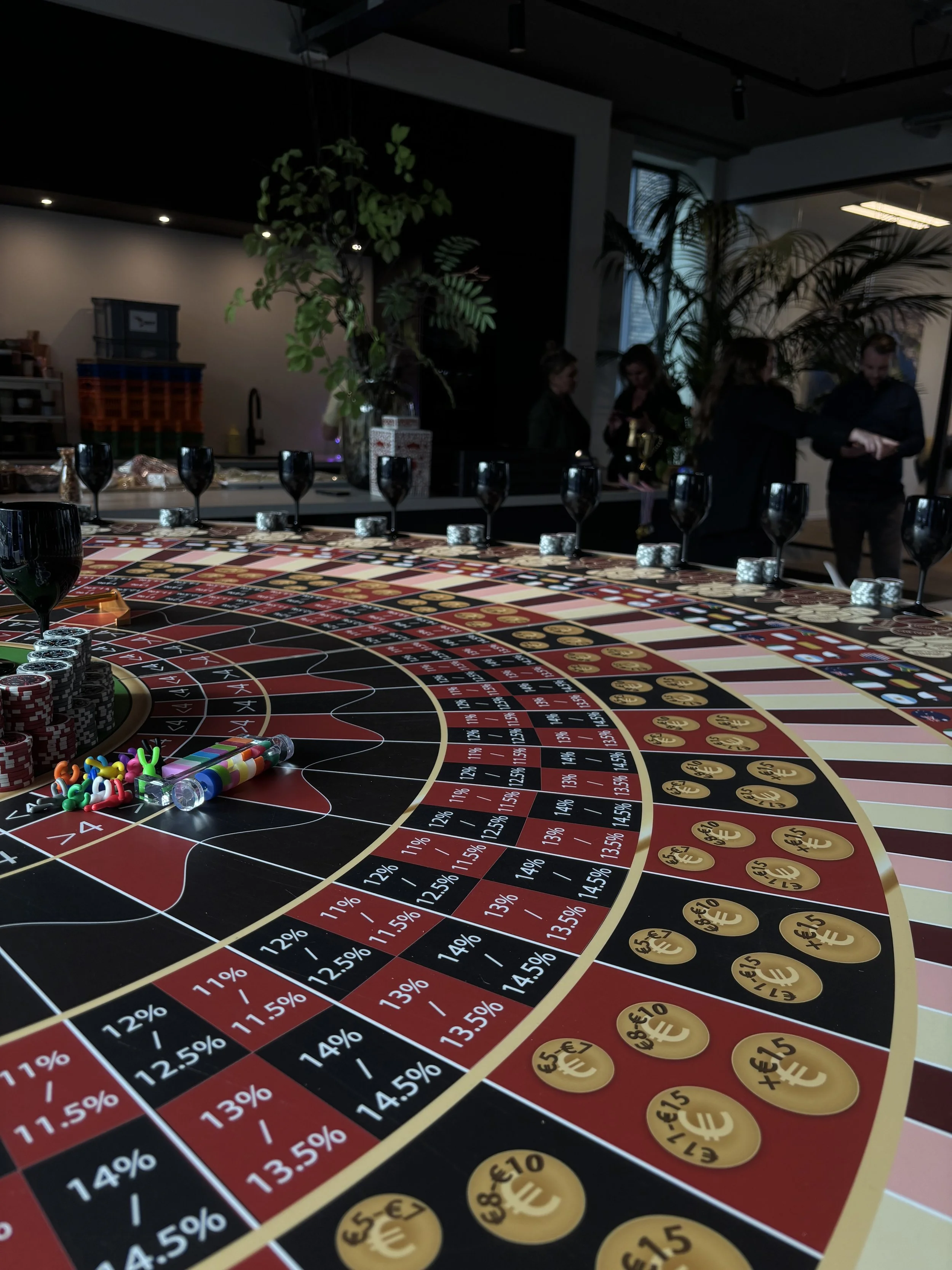 Een roulette tafel met zwart, rood, en groene vakken, muntstukken en fiches. In de achtergrond zijn mensen die aan een bar of tafel staan, met planten en ramen.