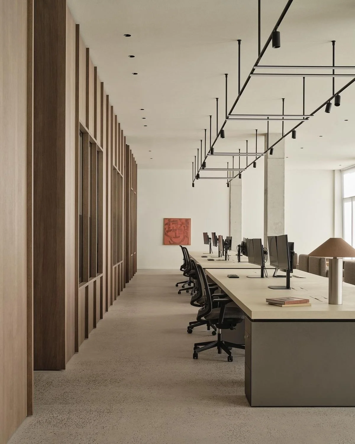 An empty modern office space with a long desk, multiple computer monitors, and black office chairs. There are wood-paneled walls on the left and a large window on the right, with minimalist ceiling lighting fixtures and a red abstract painting on the wall.